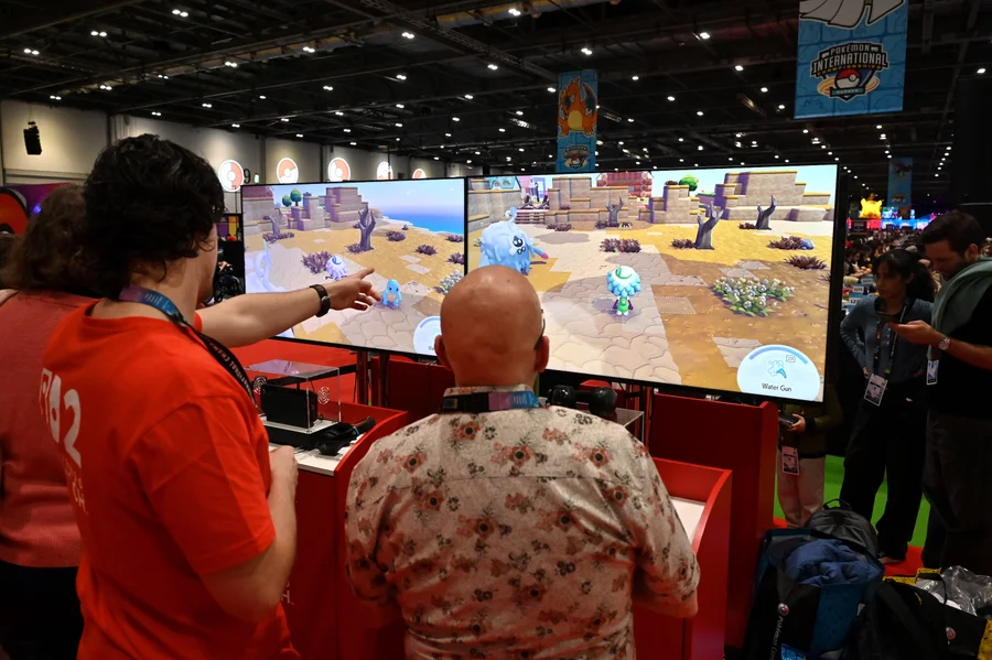 people play a video game on two large monitors. the characters on the screen are walking through a rocky landscape near an ocean