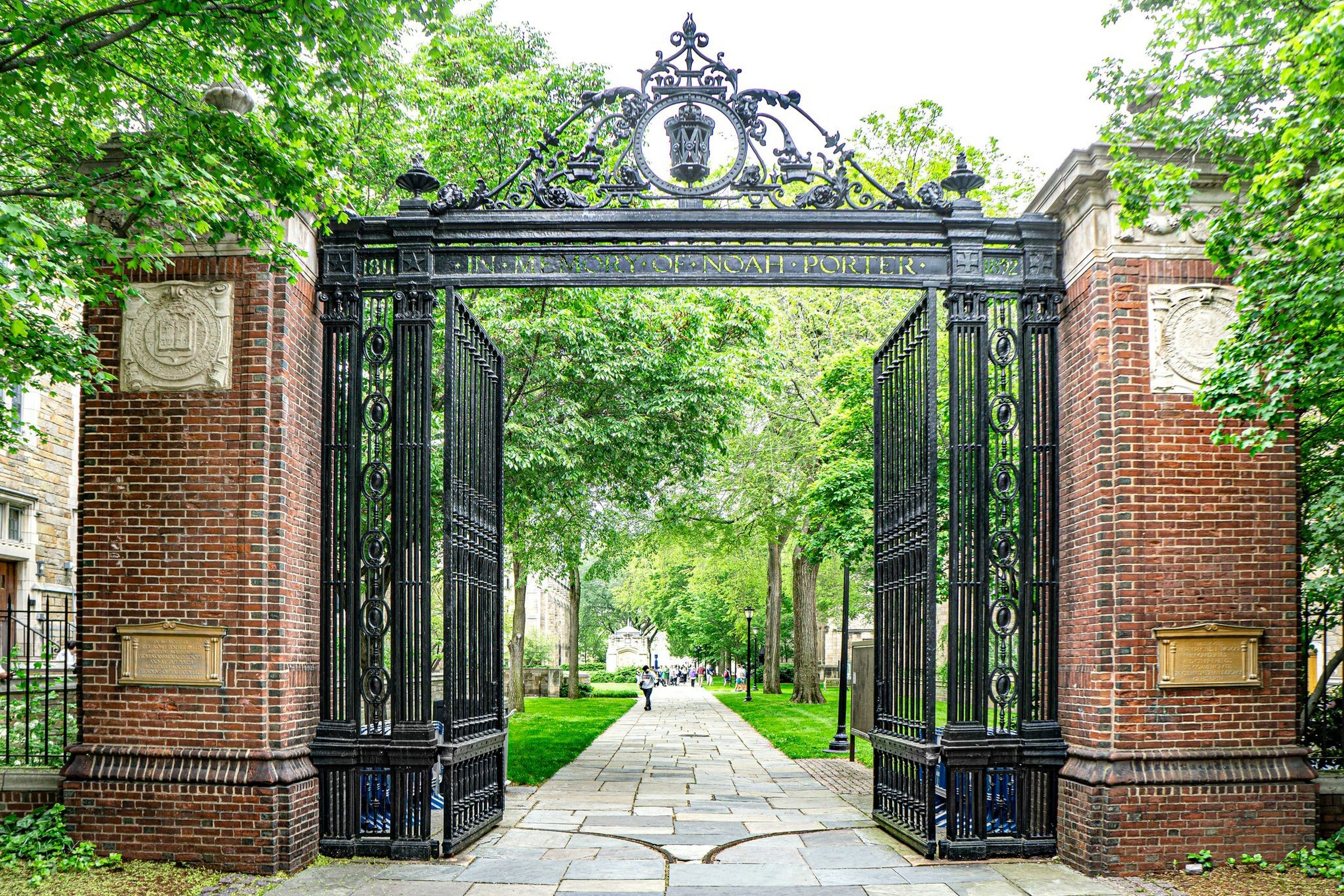 An ornate black iron gate with brick pillars stands open, leading to a tree-lined stone pathway with people walking in the distance.