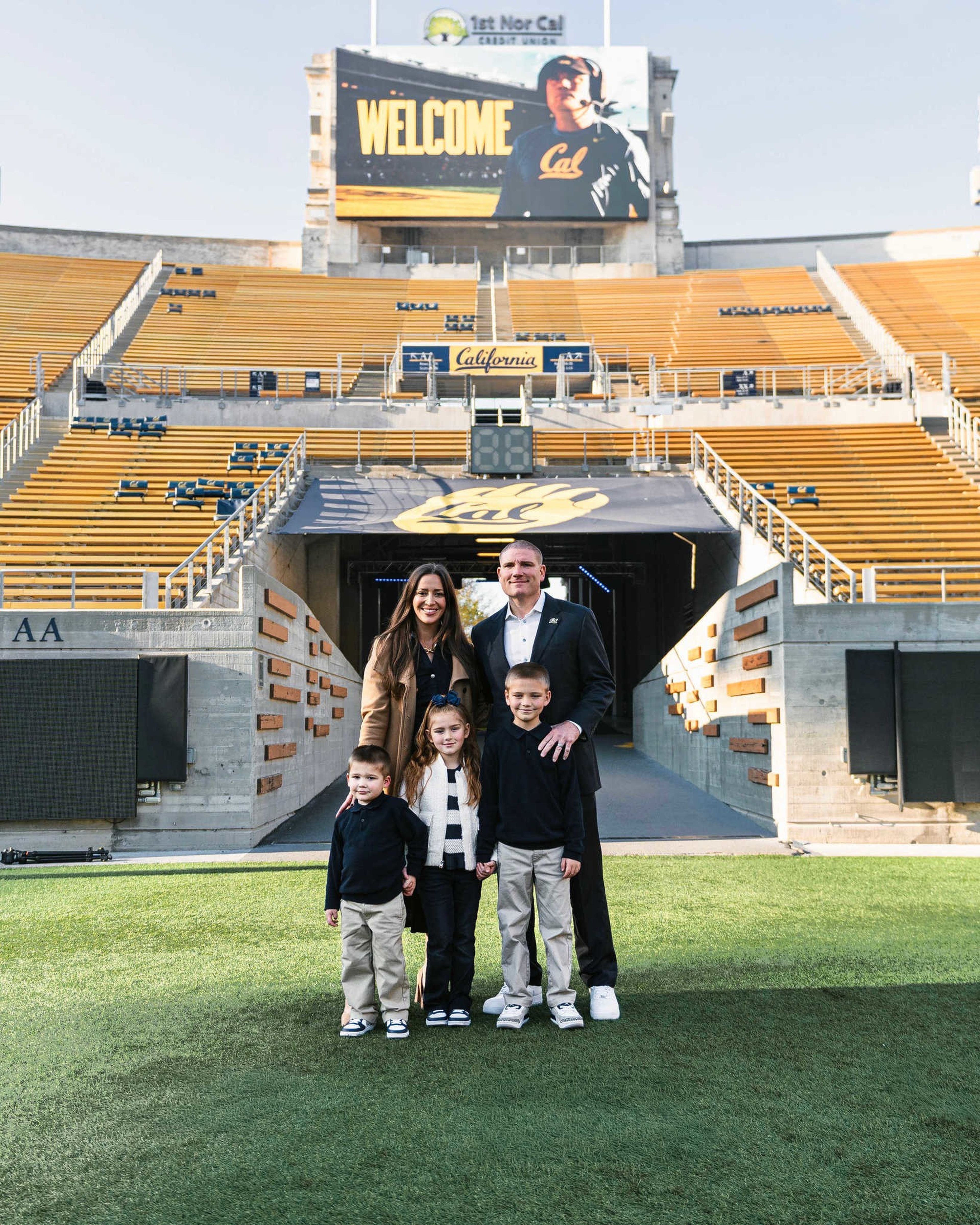 A family of five stands on a football field in front of an empty stadium tunnel with “Cal” logo and a large screen displaying “WELCOME.”