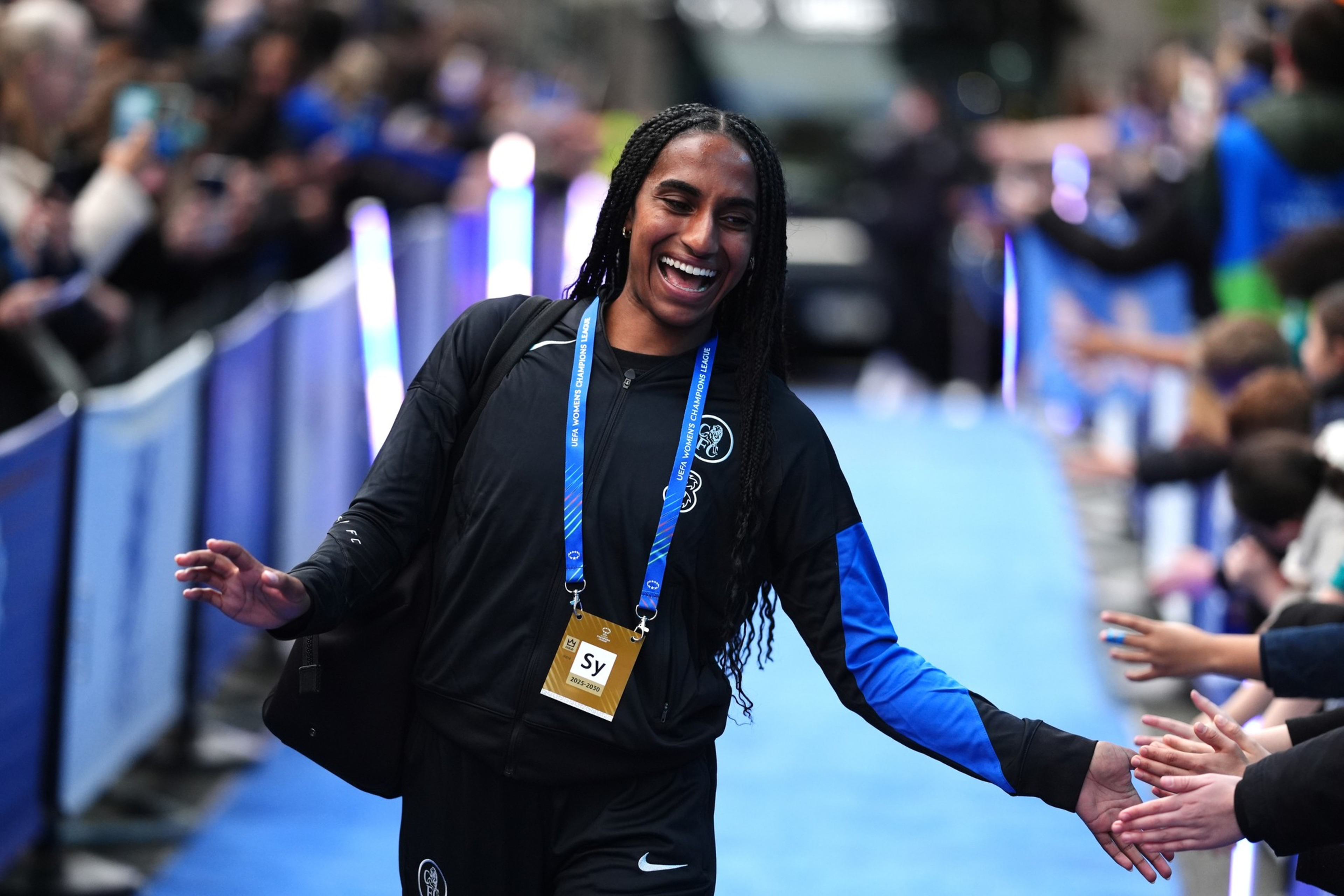 A smiling person with braided hair wearing a black and blue tracksuit high-fives a crowd along a blue carpeted pathway.