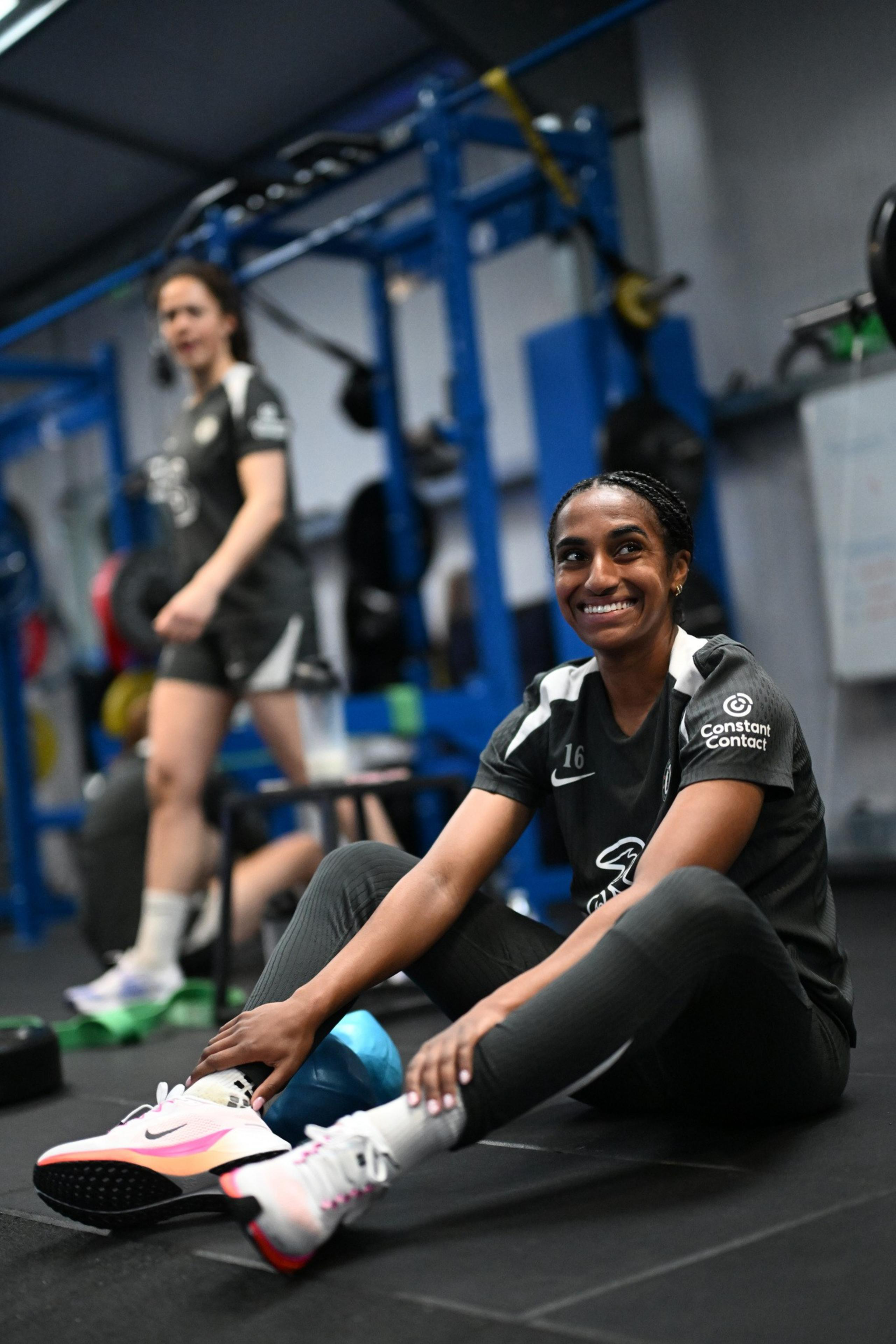 A woman in athletic gear sits on the floor smiling, stretching her legs in a gym with another woman standing and exercise equipment in the background.