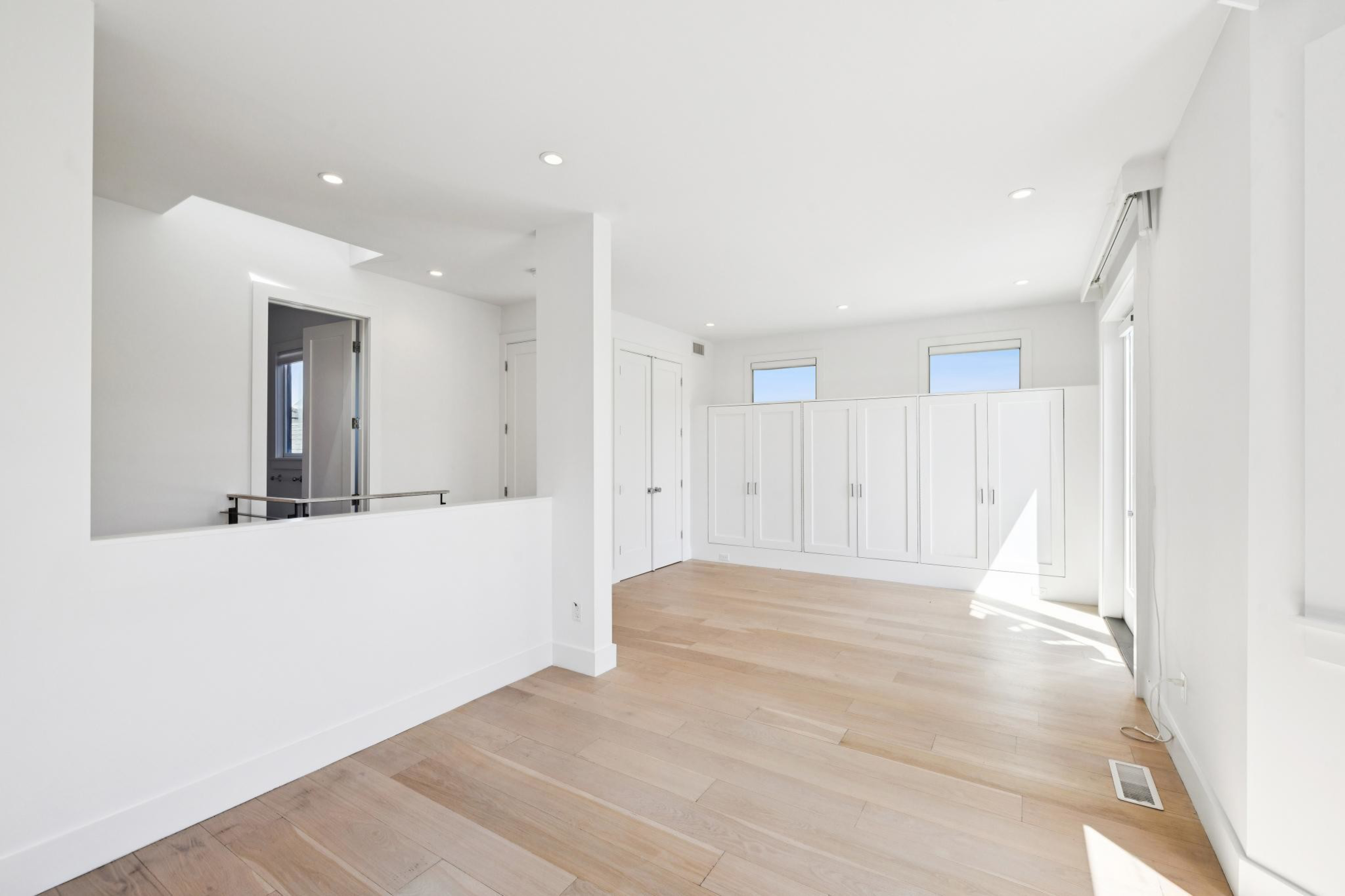 A bright, empty room with light wood floors, white walls, built-in white cabinets, two small windows, and multiple ceiling recessed lights.