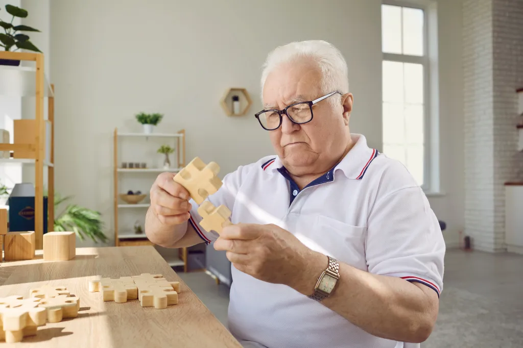 A concentrated senior man in glasses is putting together a wooden puzzle.