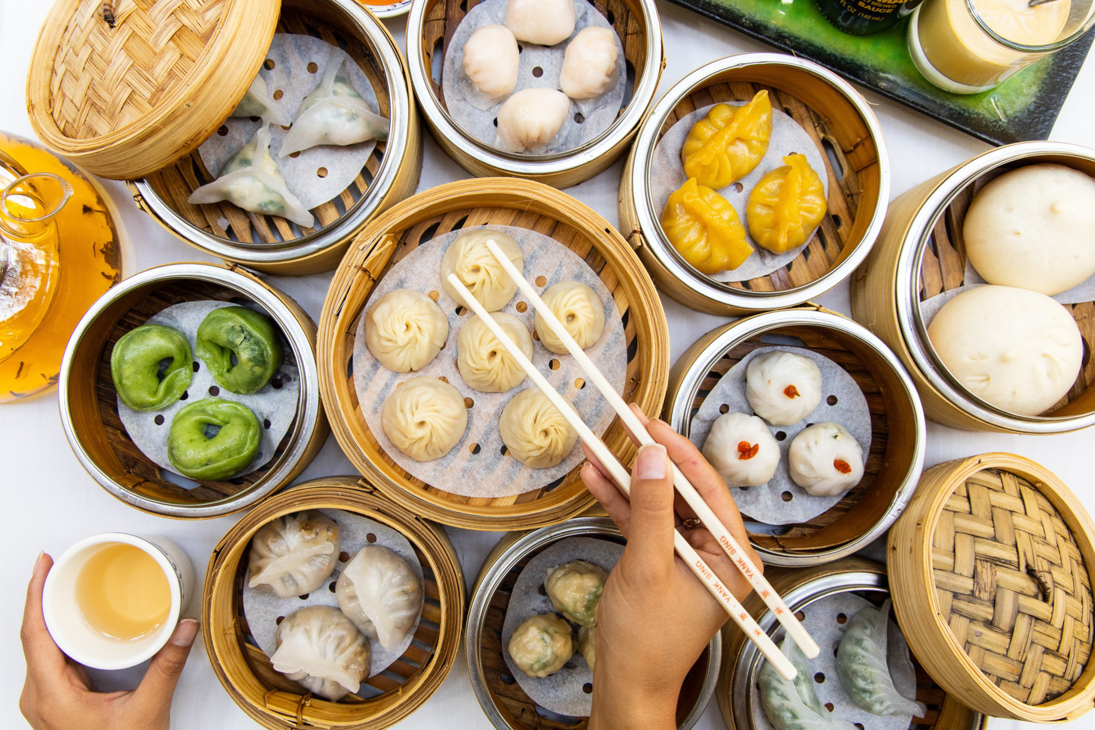 Various types of colorful dim sum, including dumplings and buns, are arranged in bamboo steamers, with hands holding chopsticks and a tea cup nearby.