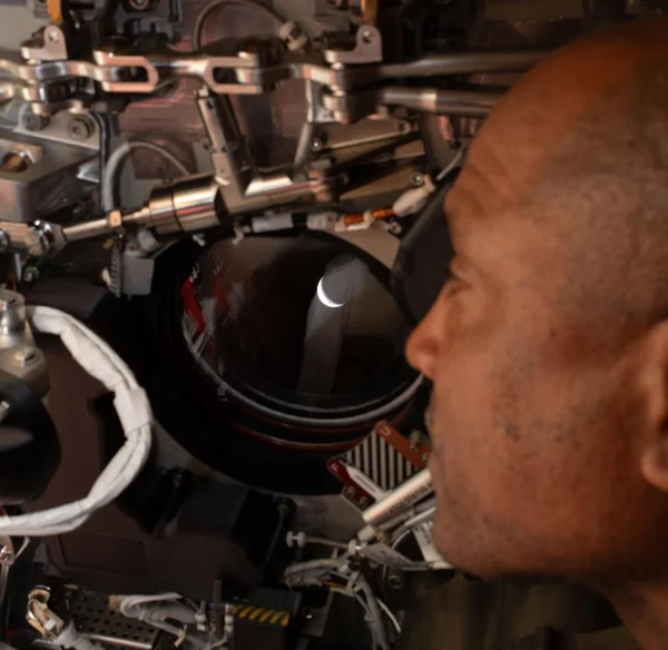 A man in a spacecraft looks out a small round window showing a bright crescent.