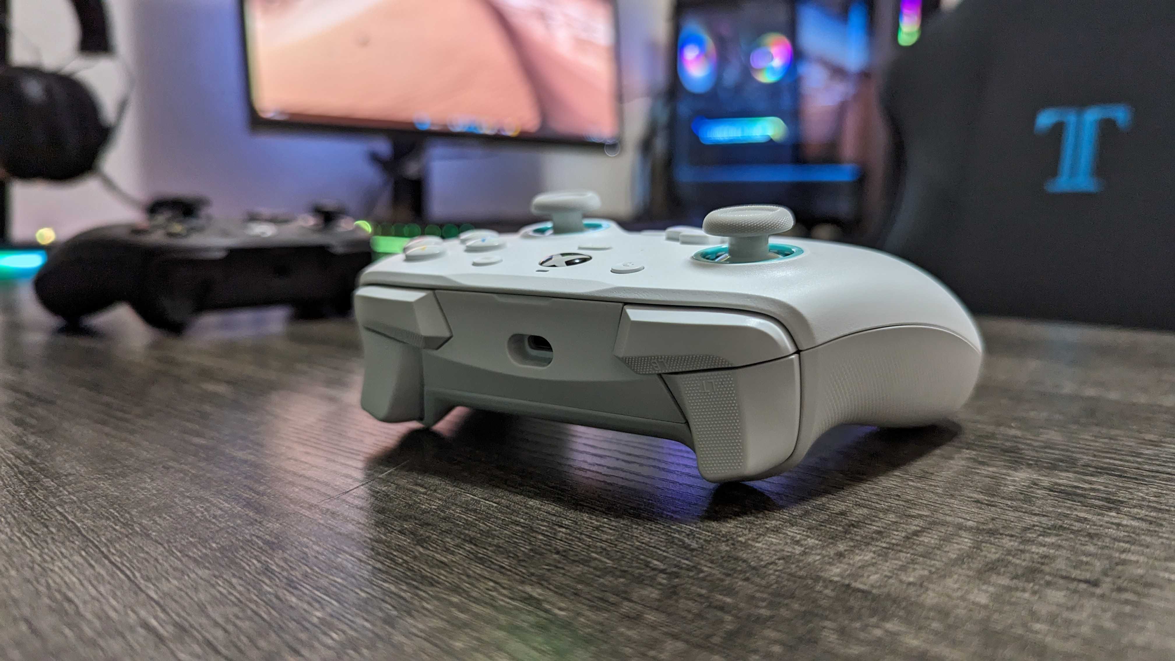 A white gaming controller rests on a wooden desk, in front of a blurred gaming PC setup with vibrant RGB lights and a monitor. Mood is tech-focused.