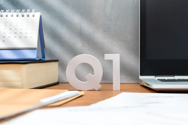 Letters Q1 on a wooden desk alongside a laptop, a book, and a desk calendar.