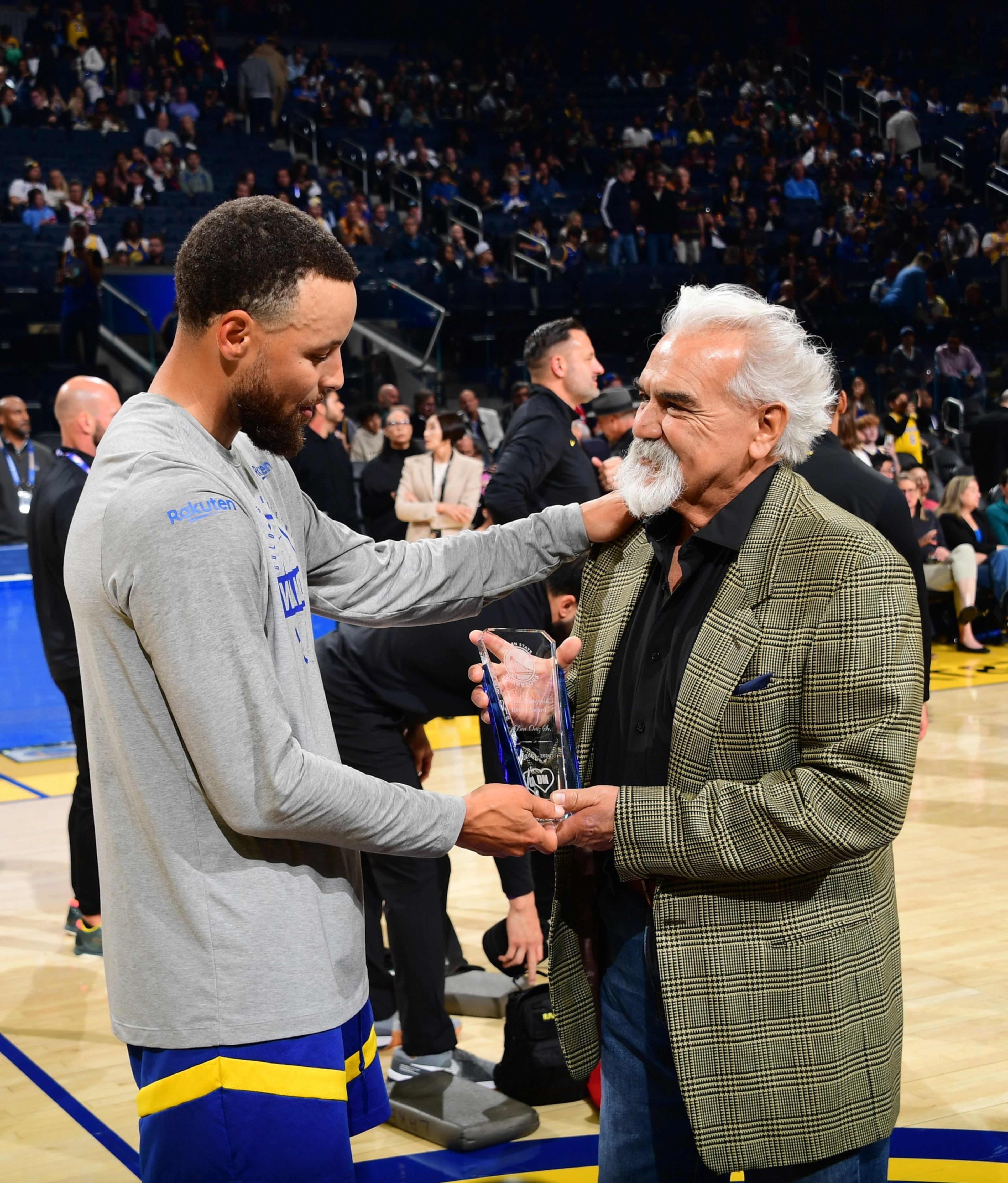 A basketball player in gray and blue presents an award and gently touches the shoulder of an elderly man with white hair and a plaid jacket.