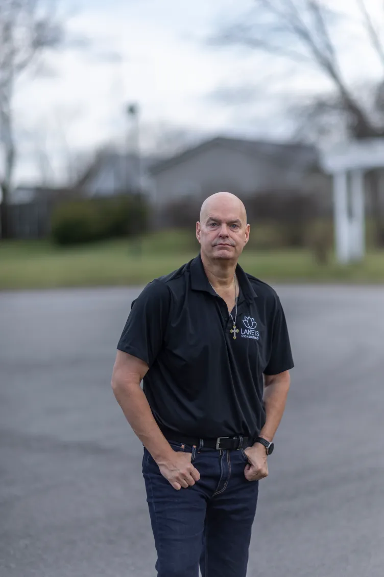 A bald man wearing dark jeans, a polo shirt and a necklace with a cross stands with his hands in his pockets. The background is a blurred road and buildings with vegetation.