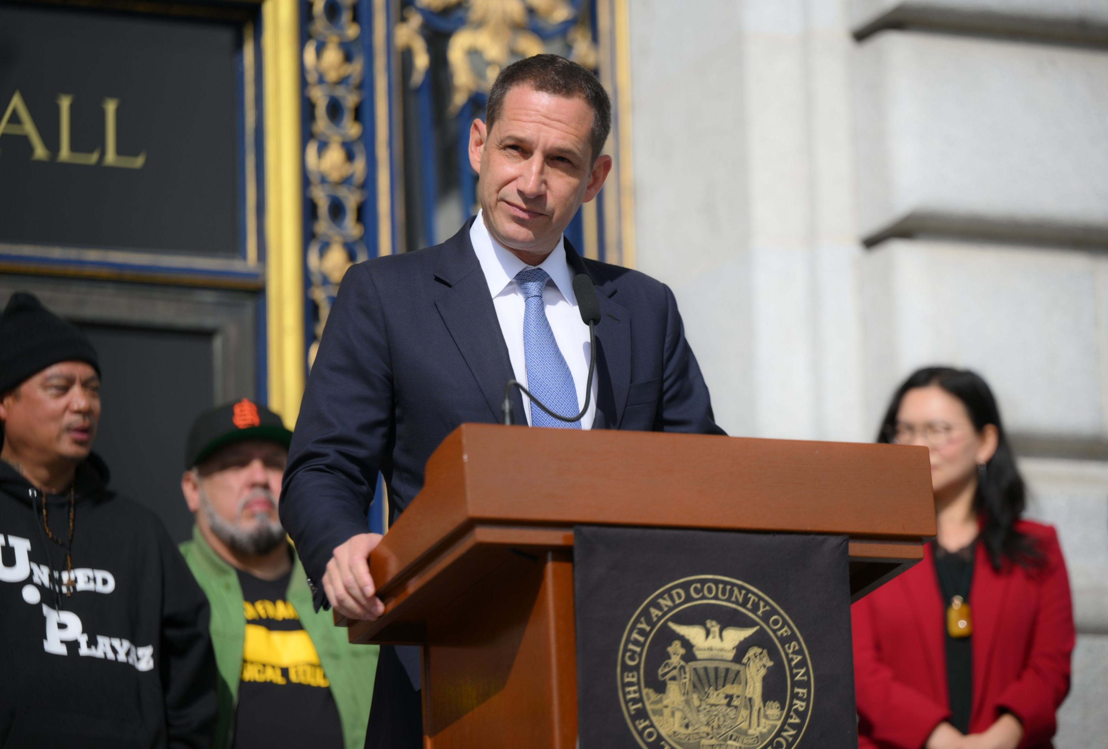 A man in a suit speaks at a podium with a San Francisco seal, while three people stand behind him in casual and business attire.