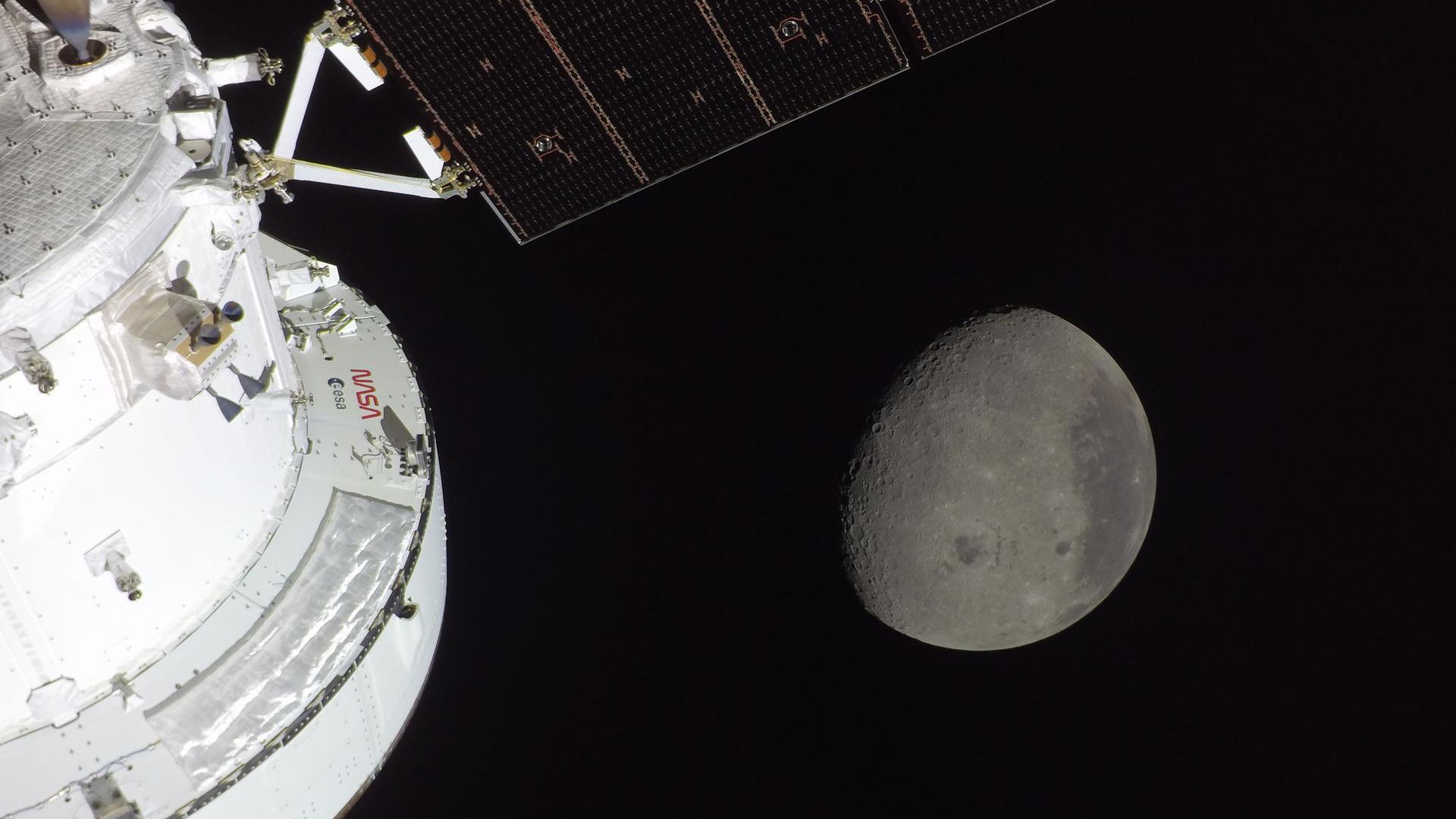 A gray sphere in the darkness of space is seen from a white spacecraft with solar panels to the left of the image. 