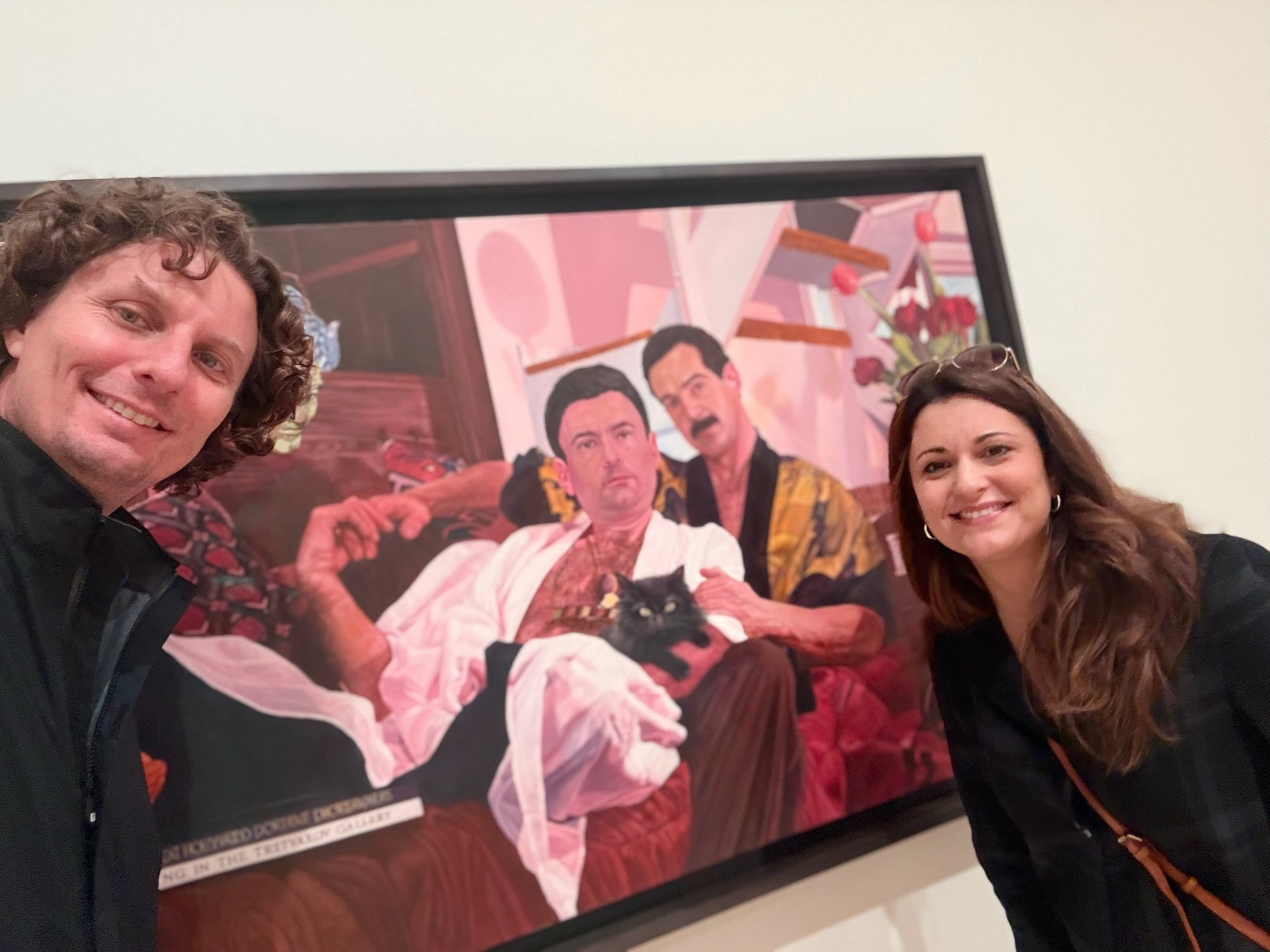 A man and a woman smile beside a framed painting of two men sitting on a couch, one holding a black cat.