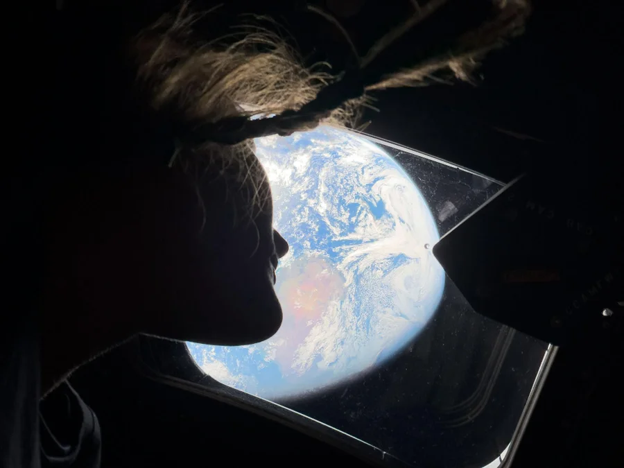Astronaut Christina Koch looks out a window of the Orion spacecraft with a pale-white earth large in the distance.