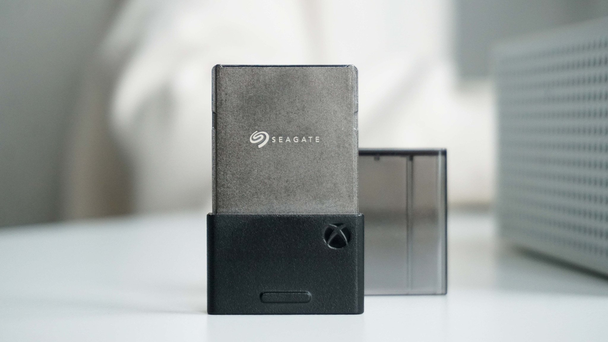 Xbox Series X|S Seagate Storage Expansion Card on a white table, inserted into a black Xbox adapter. The background is softly blurred with a light, neutral tone.