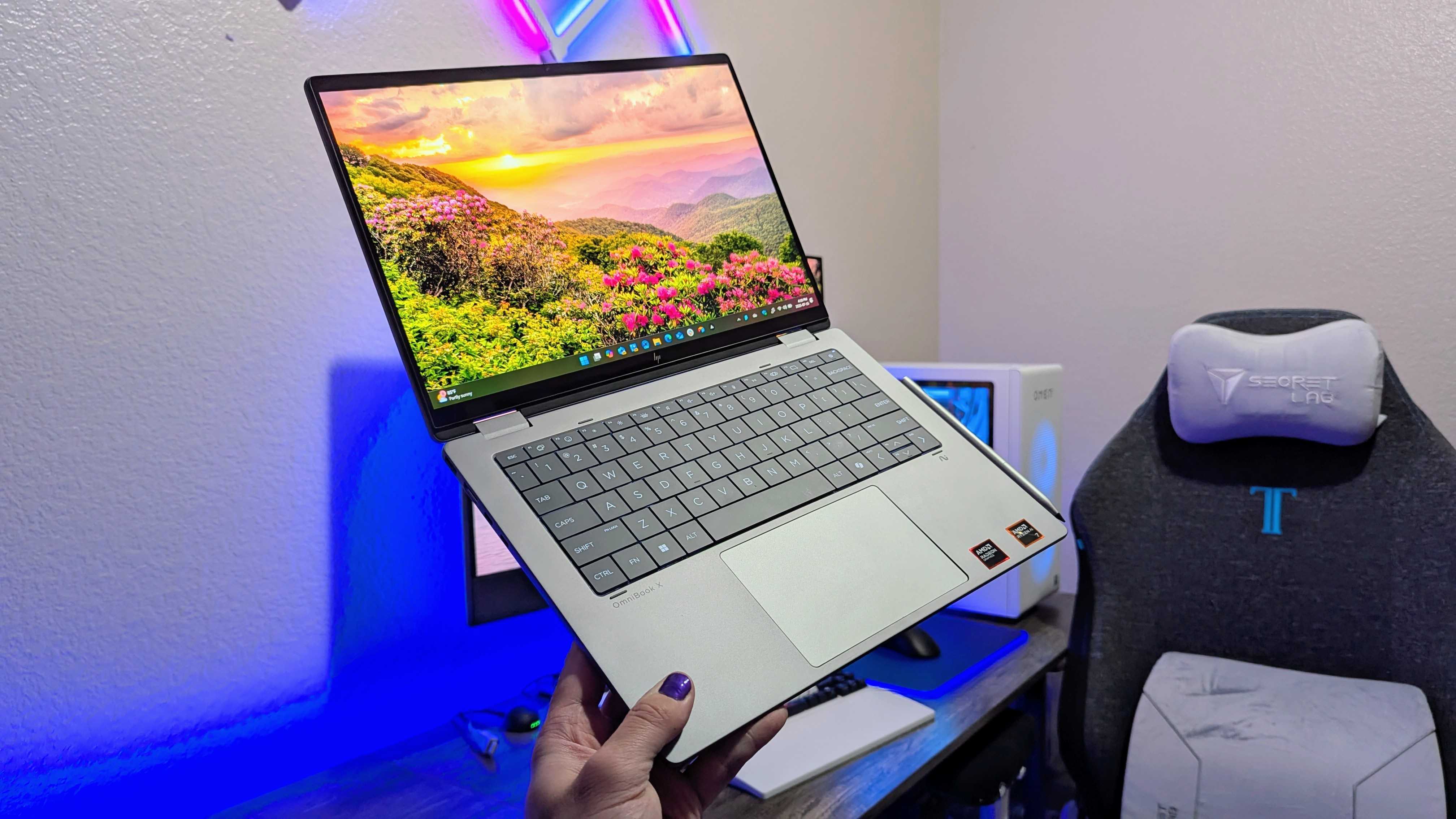 A person holds a sleek silver laptop with a vivid screen displaying a sunrise over a flower-filled landscape. A gaming chair and PC setup are in the background.