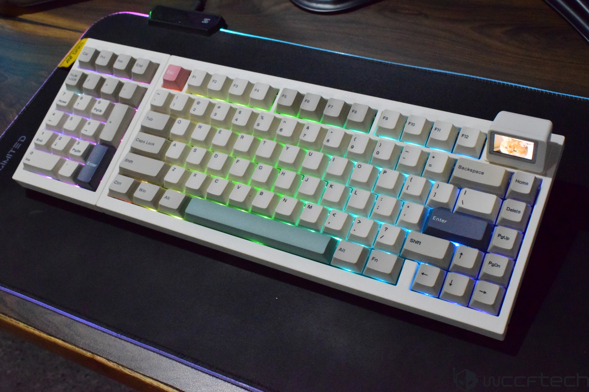 A retro-style mechanical keyboard with RGB backlighting and a small color display in the top right corner, showing an image of a cat, placed on a dark mouse pad.