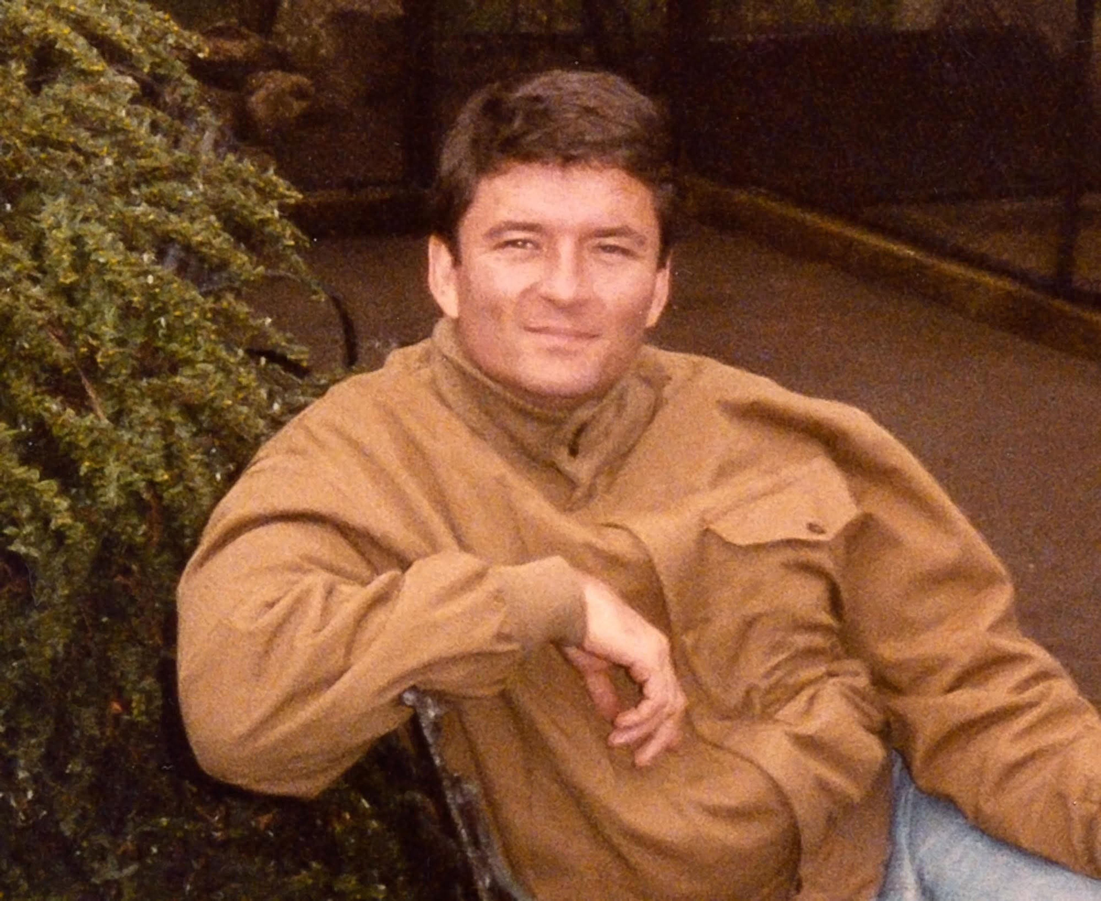 A man with short dark hair wears a brown jacket and light blue jeans, leaning casually against a tree covered in greenery.