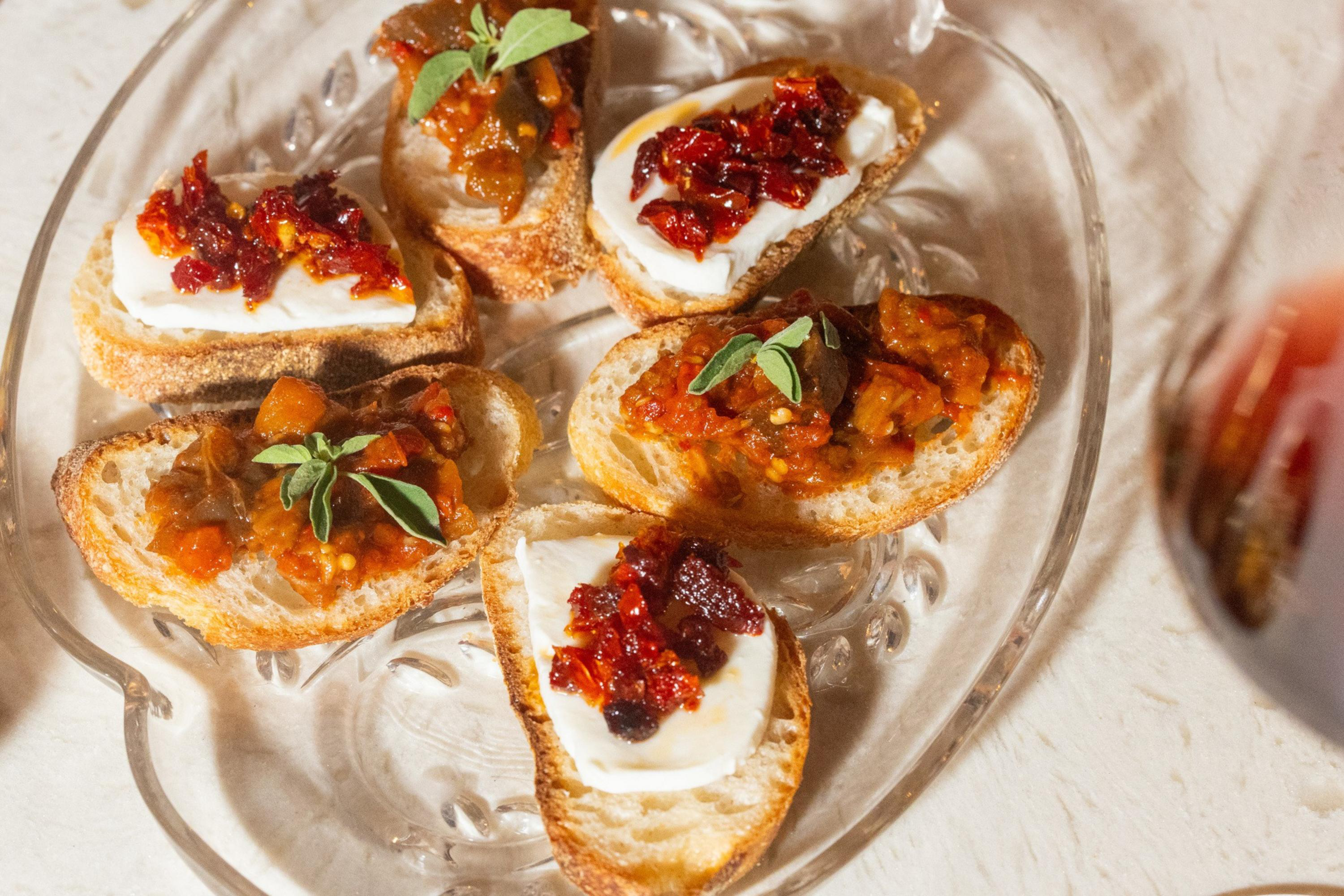 Six toasted bread slices topped with white cheese, red pepper spread, tomato relish, and fresh green herbs arranged on a clear glass plate.