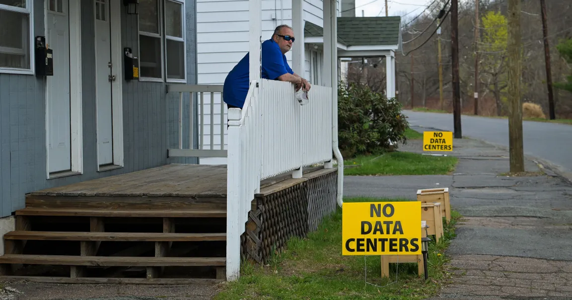 A man wearing a blue shirt and sunglasses leans on a white wooden porch railing, holding a clear plastic cup. The porch is attached to a house with gray and white siding. In the foreground and background, there are bright yellow signs with black text that read "NO DATA CENTERS." The scene is set along a quiet street with some trees and utility poles visible.