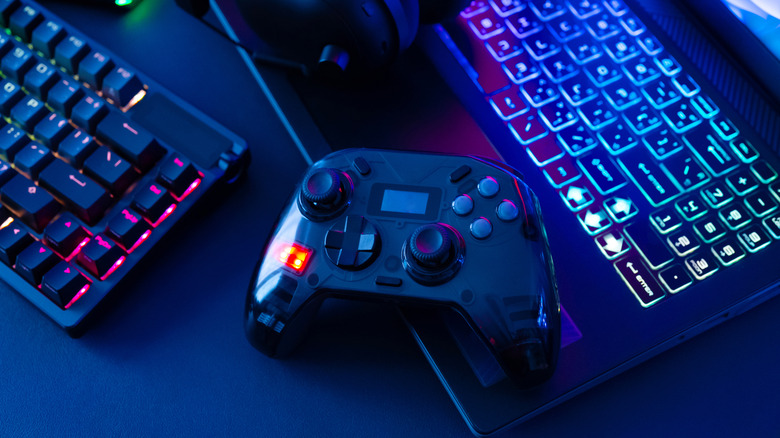 A game controller lying alongside a laptop and a keyboard with RGB lights