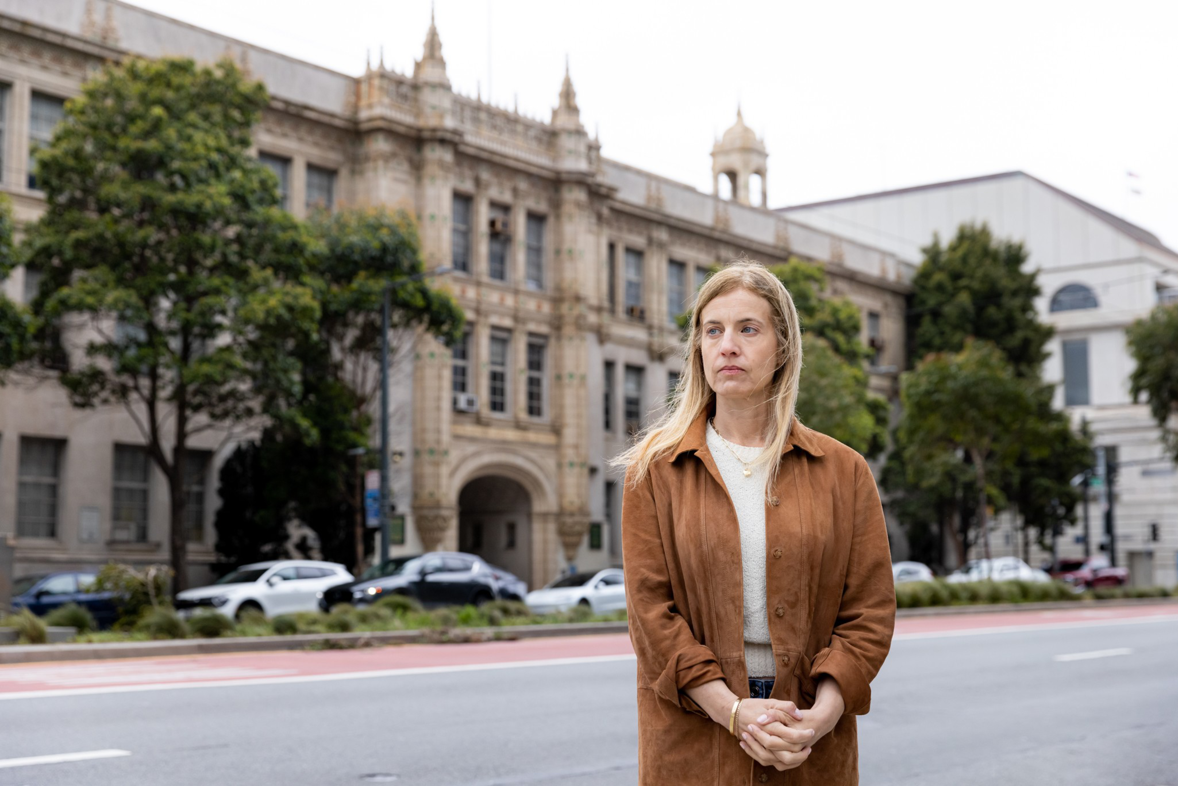 A woman with long blonde hair, wearing a brown jacket and white shirt, stands with clasped hands on a city street in front of a large historic building.