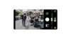 A screenshot of a mobile phone camera app in use, showing a group of four people standing on a city street. The camera is in "Photo" mode. White square outlines are overlaid on each person's face, indicating that the Guided Frame feature is actively detecting them for an accessible photo-taking experience. The overall scene is bright, with parked cars and buildings in the background.