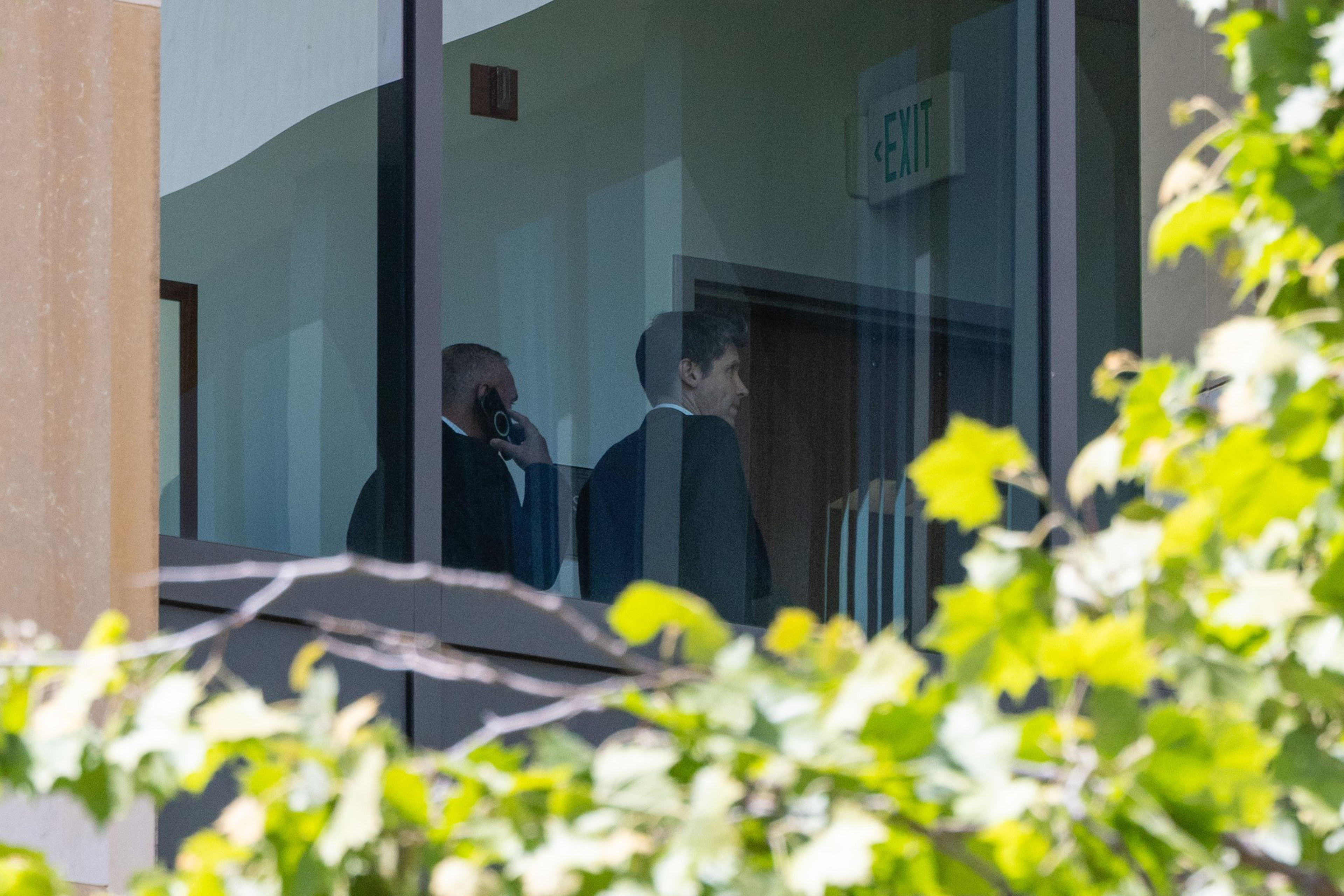 Two men in suits are seen through a glass window, one talking on a phone, standing near a door with an illuminated “EXIT” sign above it.