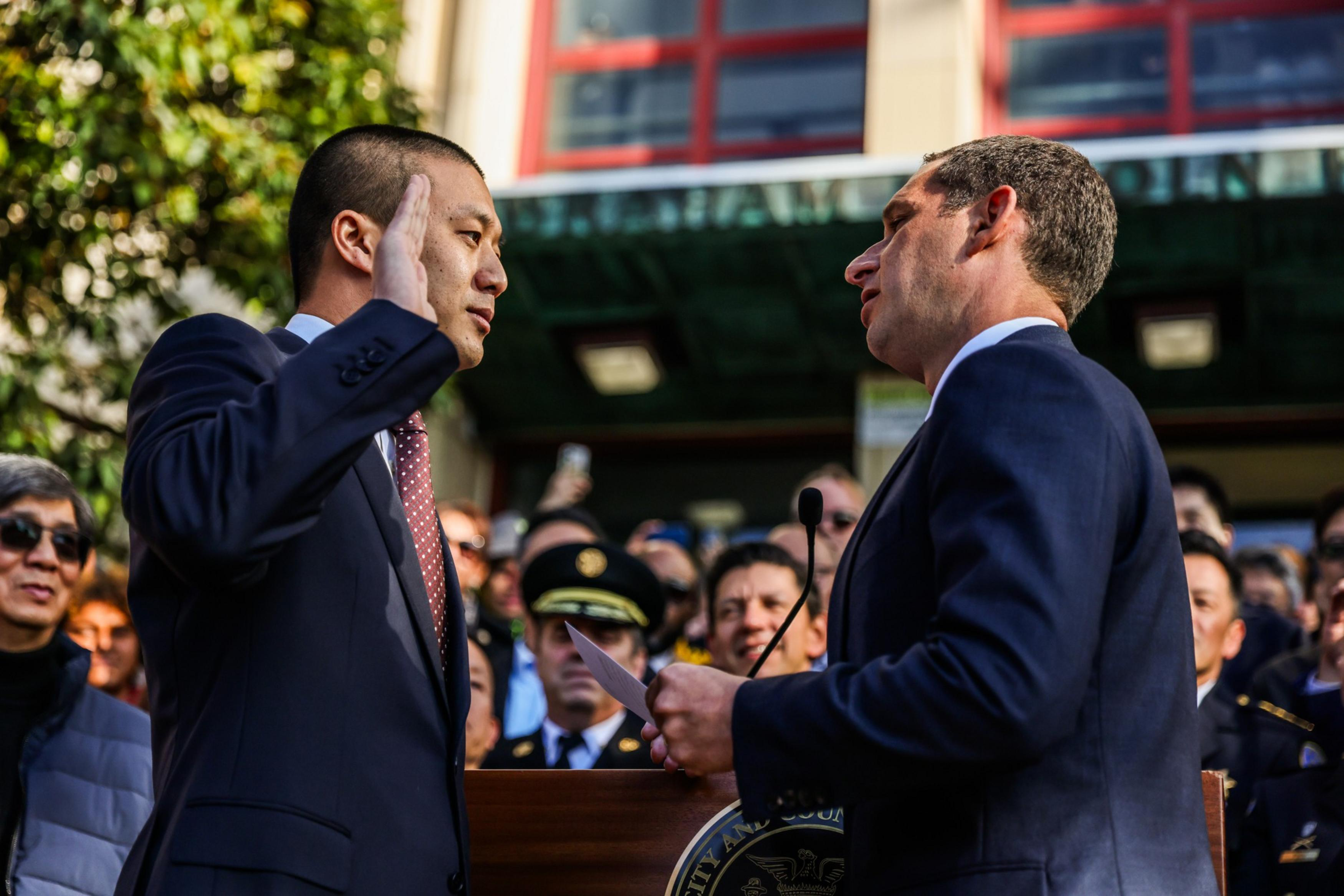 A man in a suit raises his right hand while another man in a suit holds a paper, with a crowd and police officers watching behind them.