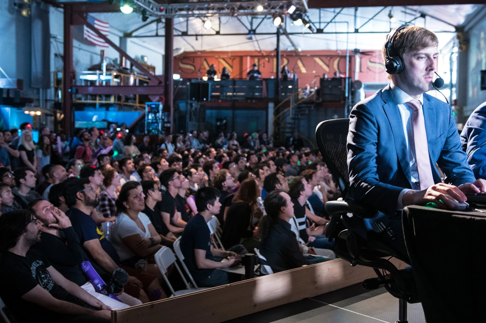 Person on stage with headphones on, playing a game on a brightly lit screen that illuminates their face while a live audience sits behind them