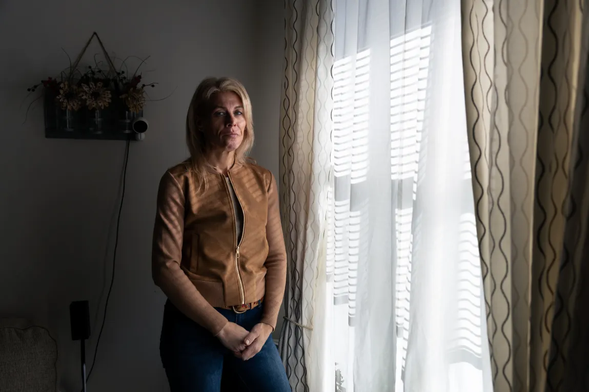 A blond woman wearing a brown jacket and jeans stands next to a window that has light streaming through the sheer white curtains. Hanging on the wall behind her is a basket of flowers.