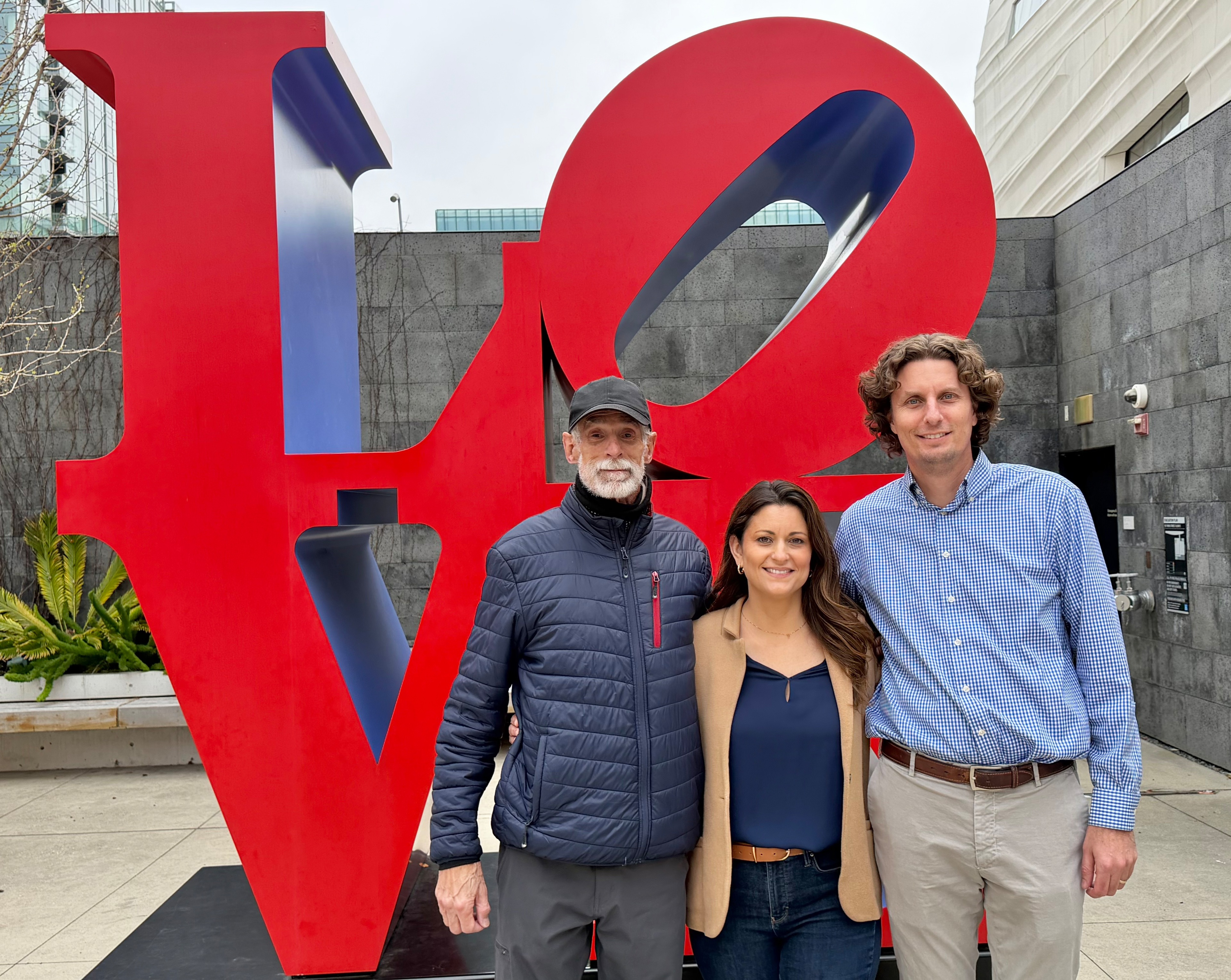 Three people stand smiling in front of a large red sculpture spelling “LOVE,” with a gray stone wall and some greenery in the background.