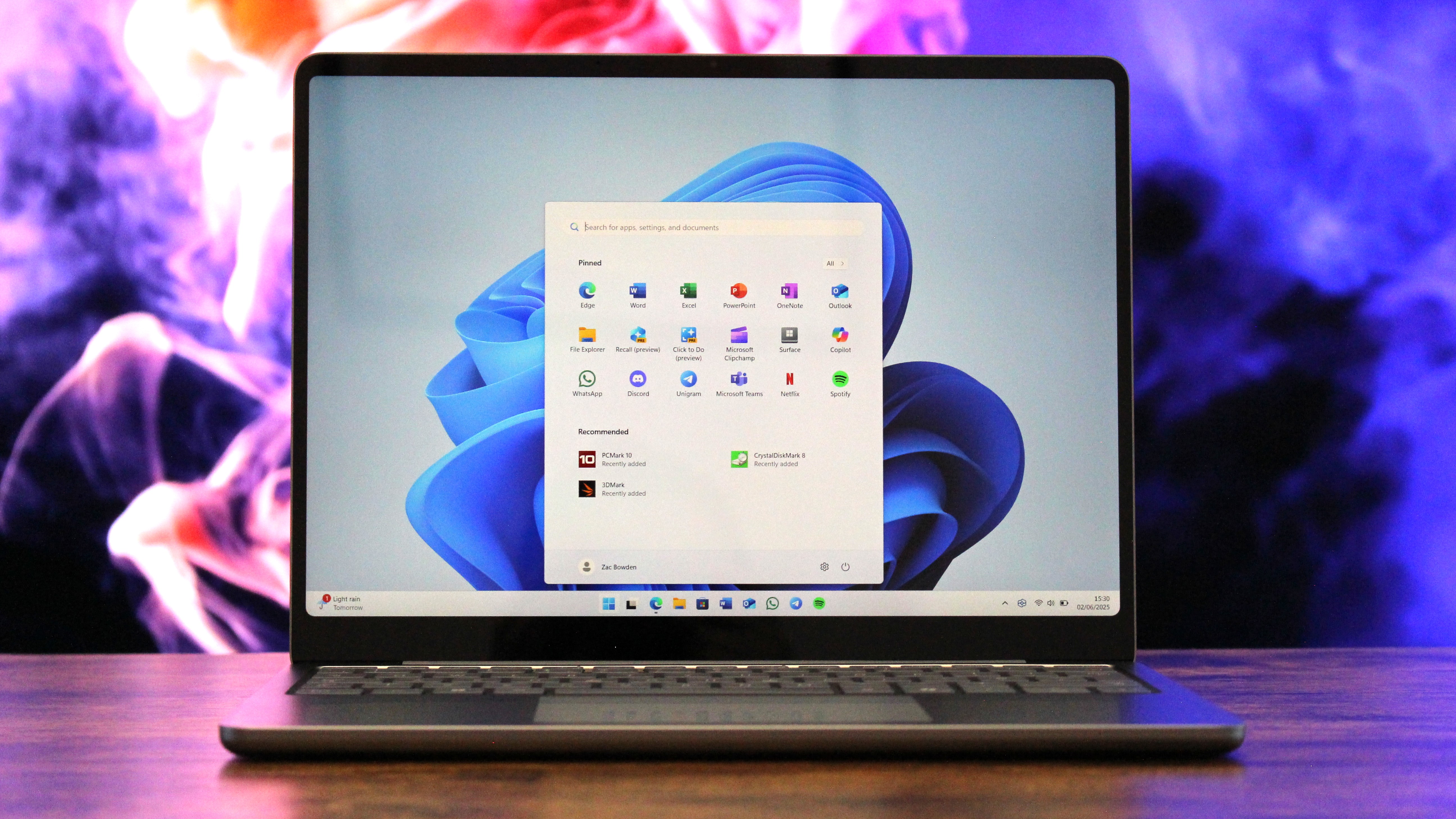 Sleek laptop with Windows 11 home screen, featuring iconic blue swirl wallpaper. The backdrop is a vibrant mix of pink, purple, and orange smoke.