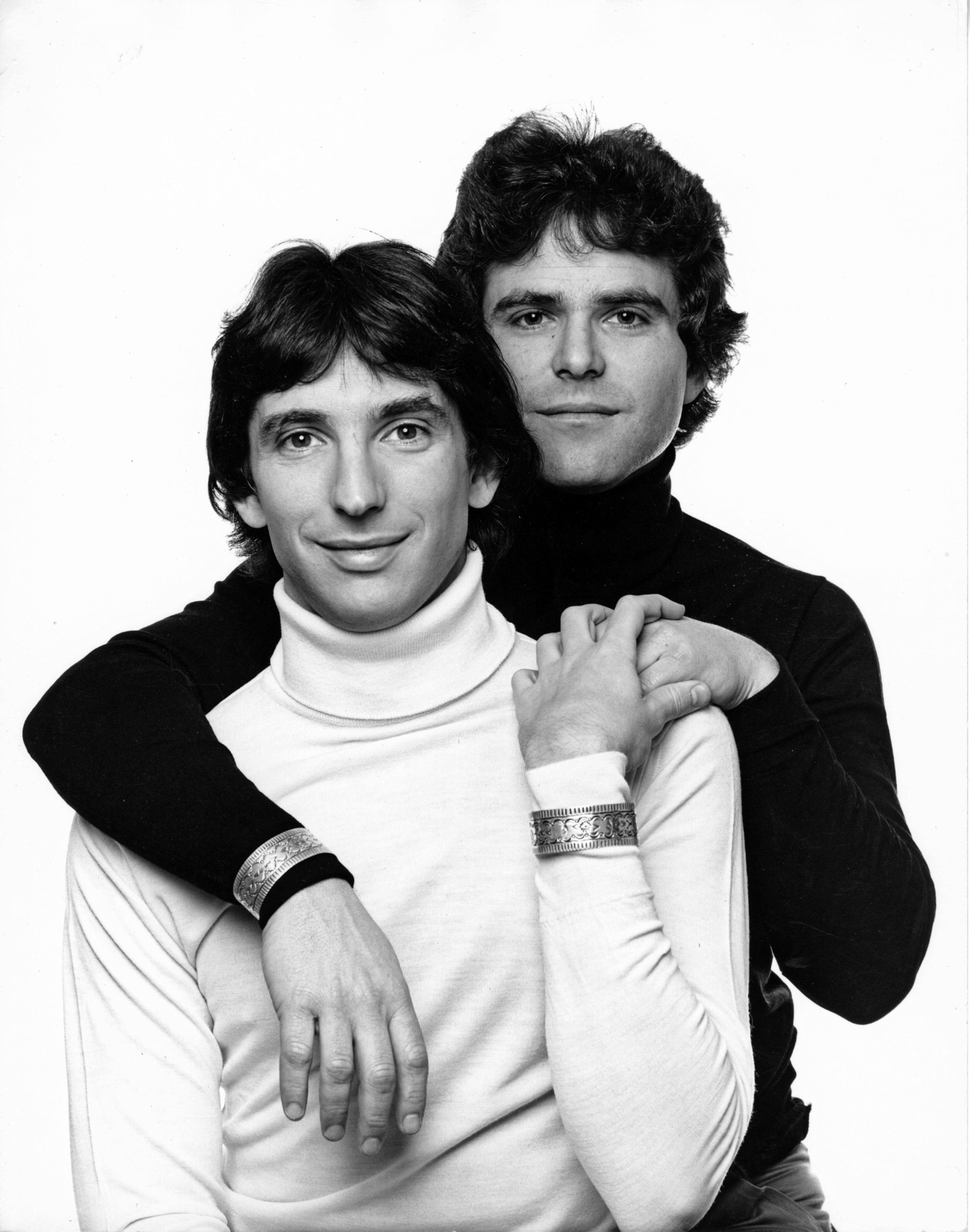 Two men pose closely, one in a black turtleneck behind the other in a white turtleneck, both wearing similar ornate bracelets and smiling softly.