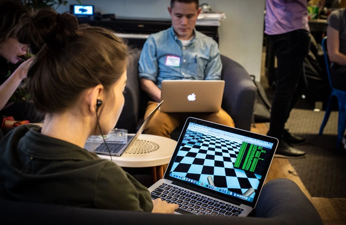 Side profile of a person sitting with earbuds in their ears, looking intently at a laptop screen of a 3D checkerboard environment