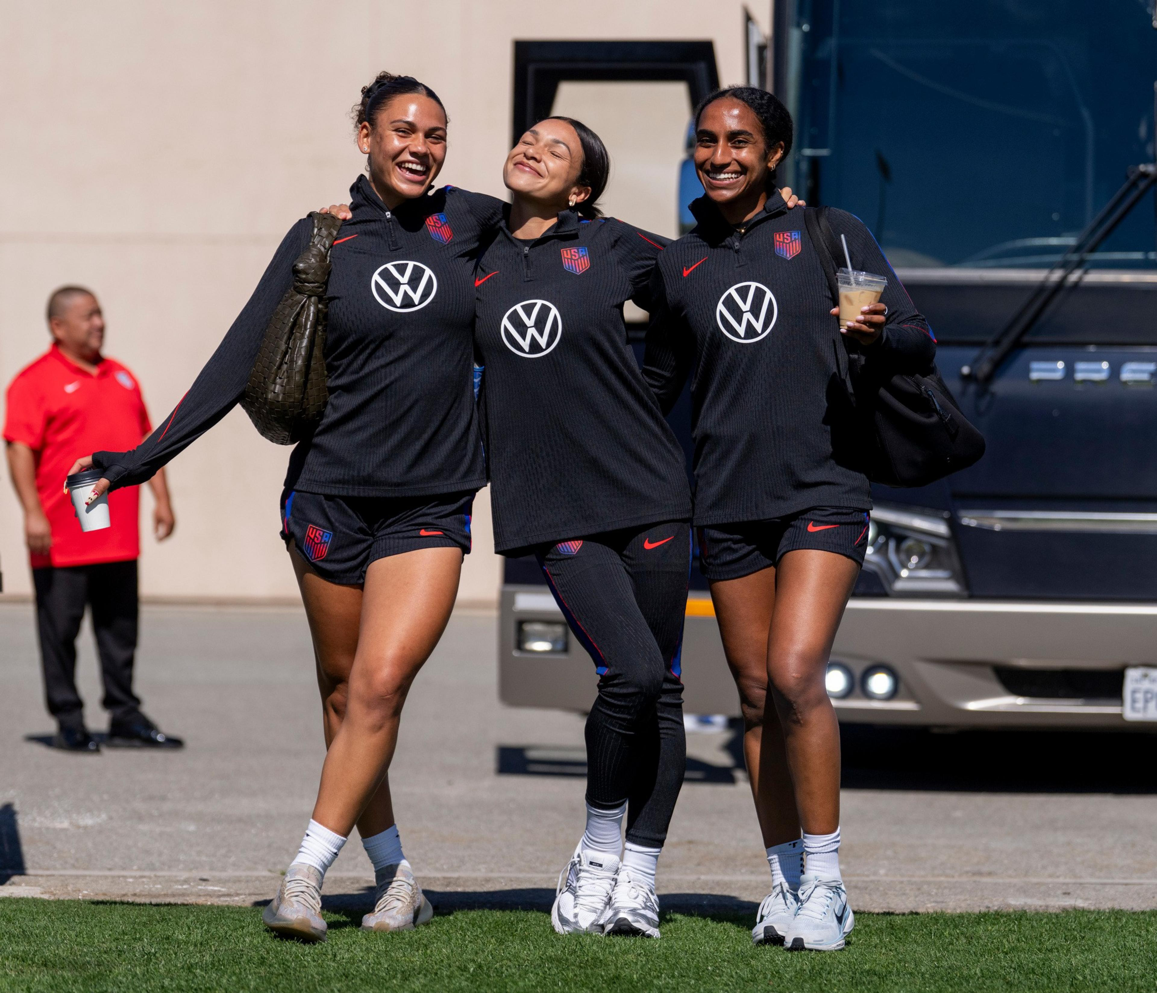 Three women in matching black sports outfits smile and walk arm-in-arm, with two holding drinks, standing outside near a bus and a man in a red shirt.