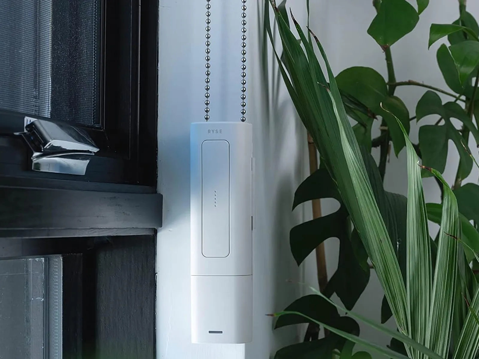 Ryse SmartShade a white rectangular box attached to a small chain dangling in a window ledge with a plant nearby