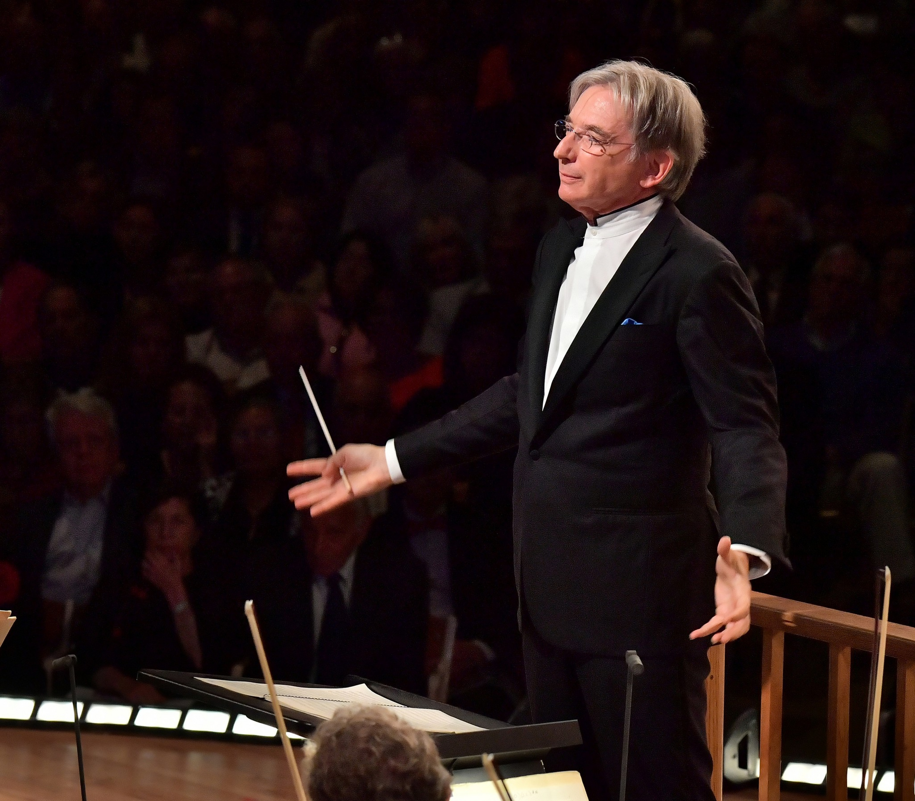 A conductor with gray hair and glasses, dressed in a black suit and white shirt, leads an orchestra with a baton in his right hand.