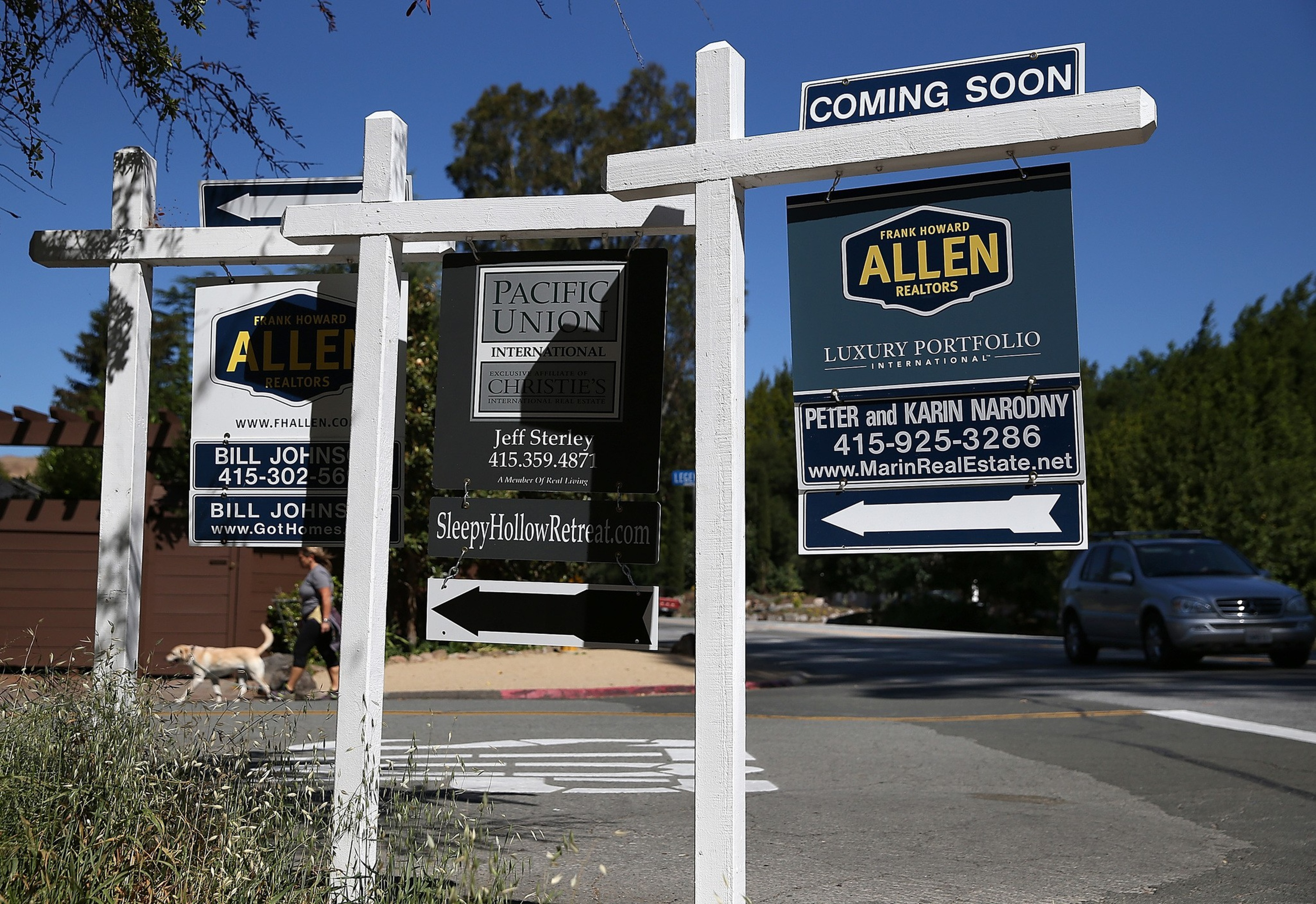 Three real estate signs hang from a white wooden post by a street corner, with one labeled “COMING SOON” and a person walking a dog in the background.