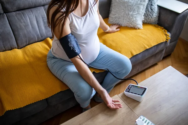 A close-up, top-down view of a pregnant woman checking her blood pressure at home. Her belly and hand are visible as she holds a digital monitor, with pregnancy supplements and vitamins placed beside her.