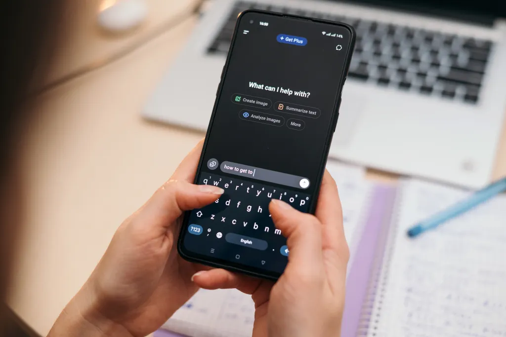 Person typing "how to get to" into an AI chat app on a smartphone, with a blurred notebook and laptop in the background.