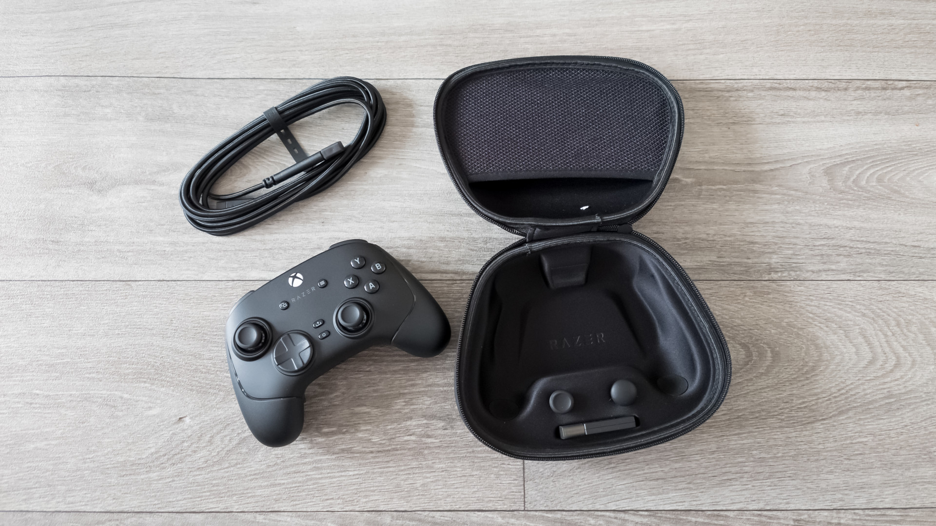 The Razer Wolverine V3 Pro wireless Xbox controller lying next to an open case and a coiled cable on a wooden floor, conveying a sleek and organized setup.