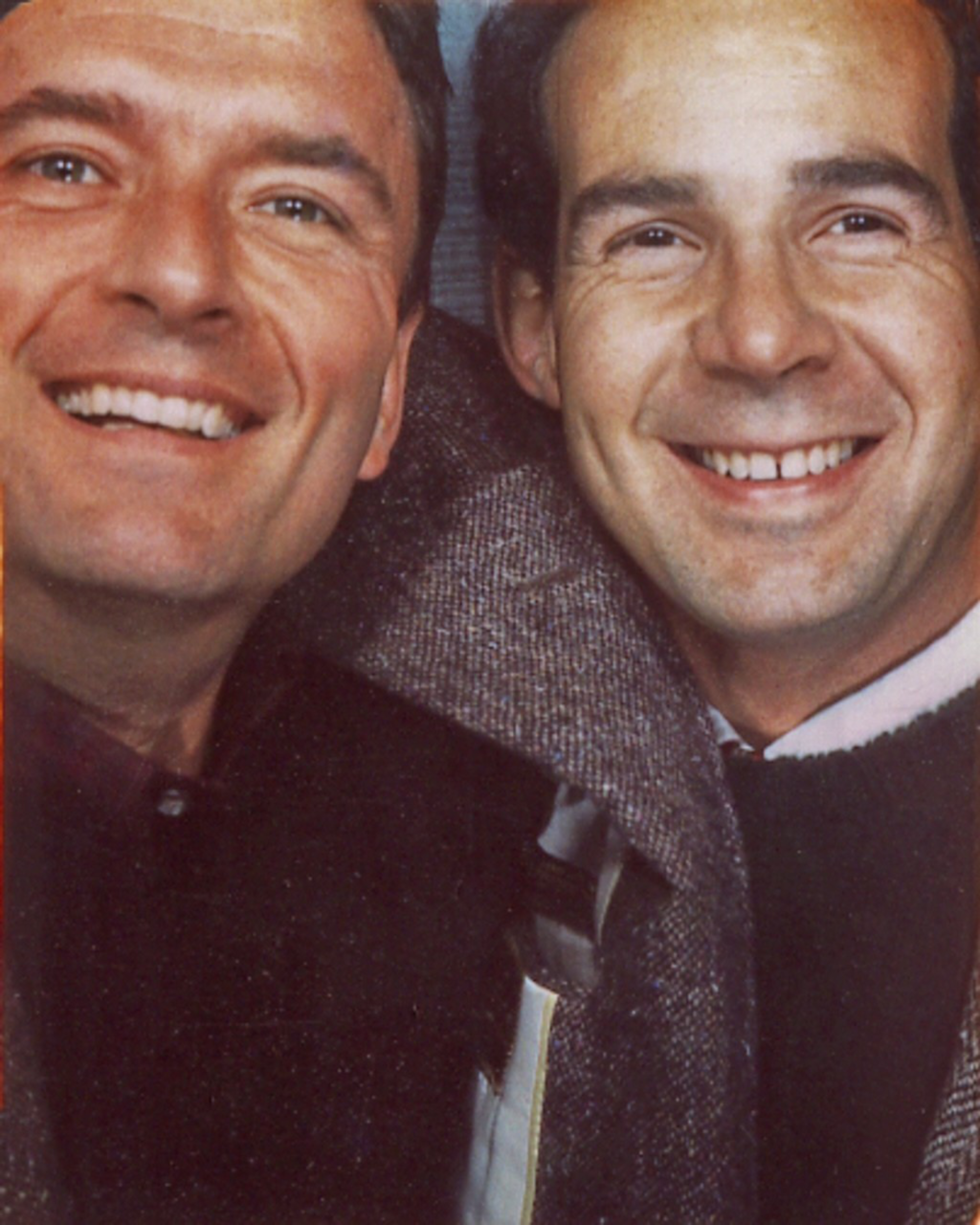Two smiling men with short dark hair are close together, wearing casual clothing including a dark sweater and a patterned jacket.