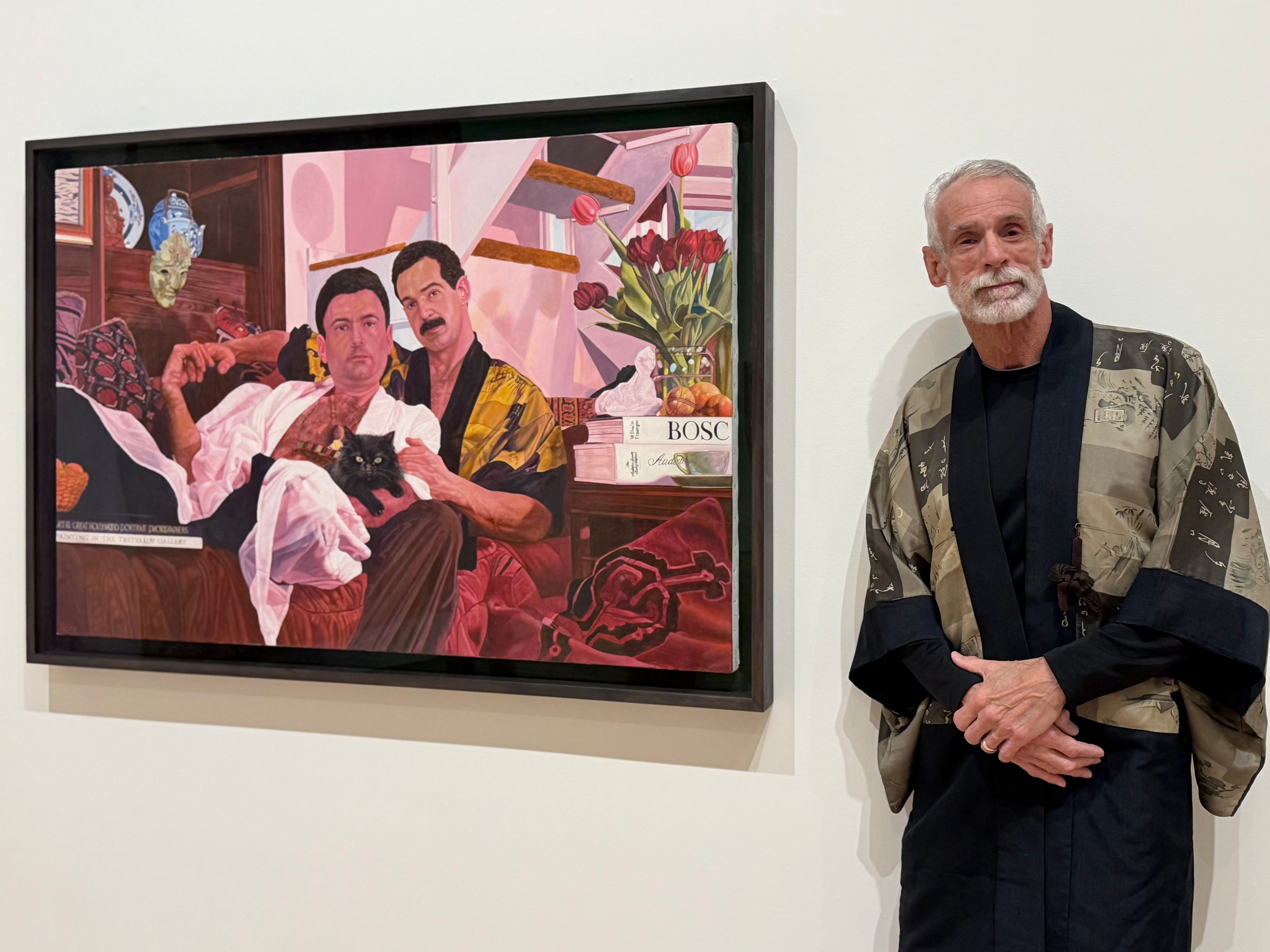 Two men seated on a couch with a black cat in one man’s lap, surrounded by books, flowers, and shelves, while an older man stands nearby wearing a patterned robe.