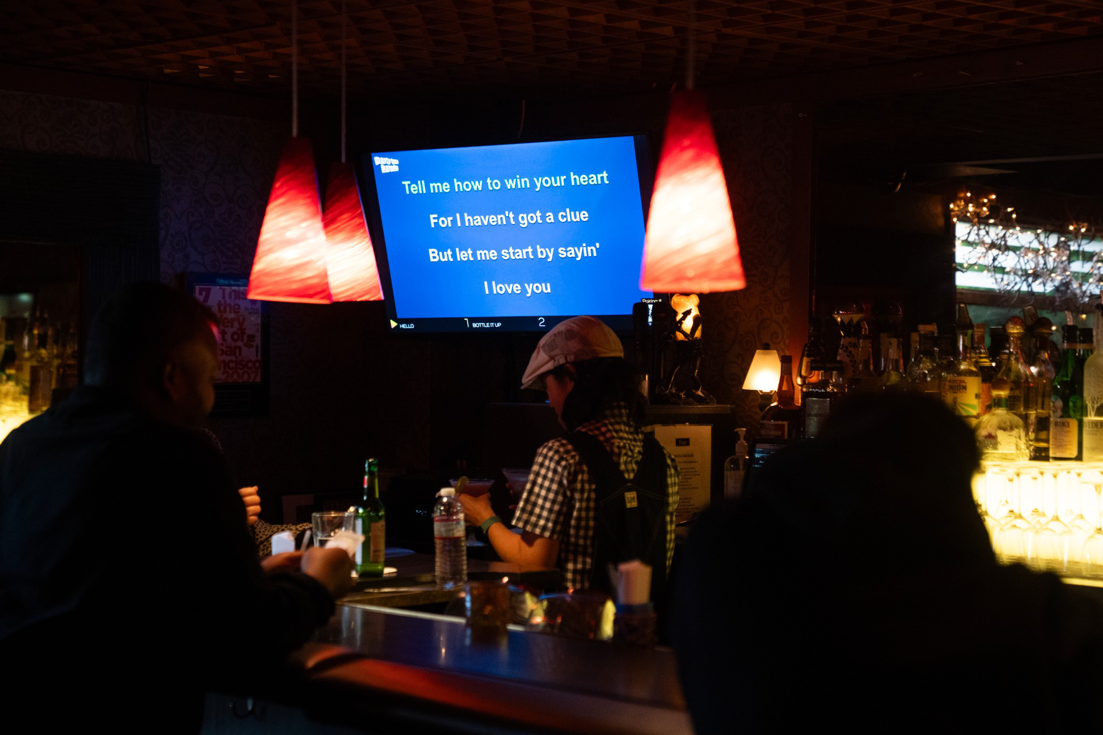 A dimly lit bar with three people sitting, red pendant lights overhead, and a karaoke screen displaying blue lyrics about love.