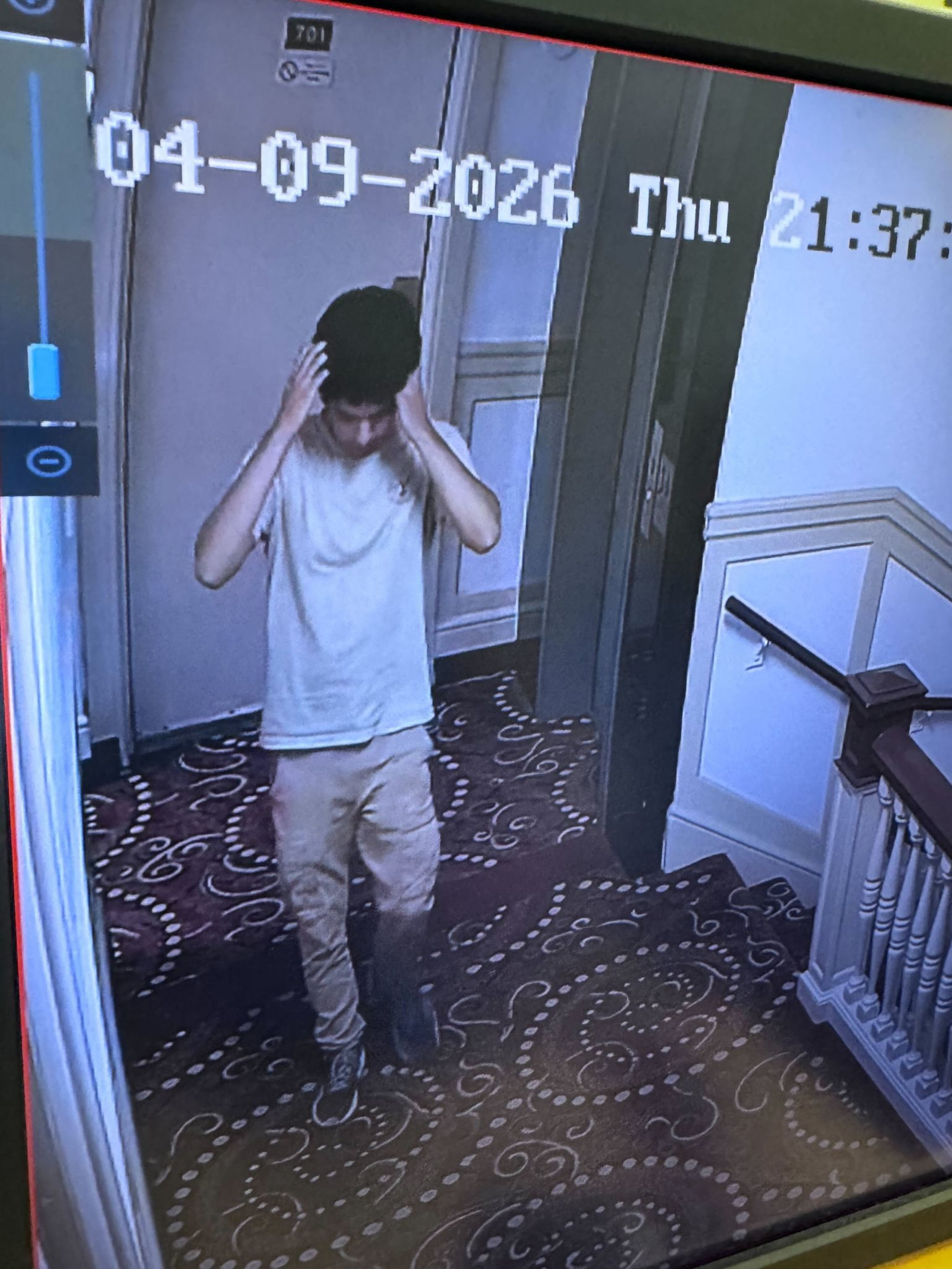 A young man in a light t-shirt and beige pants stands in a carpeted hallway with patterned carpet, holding his head with both hands.