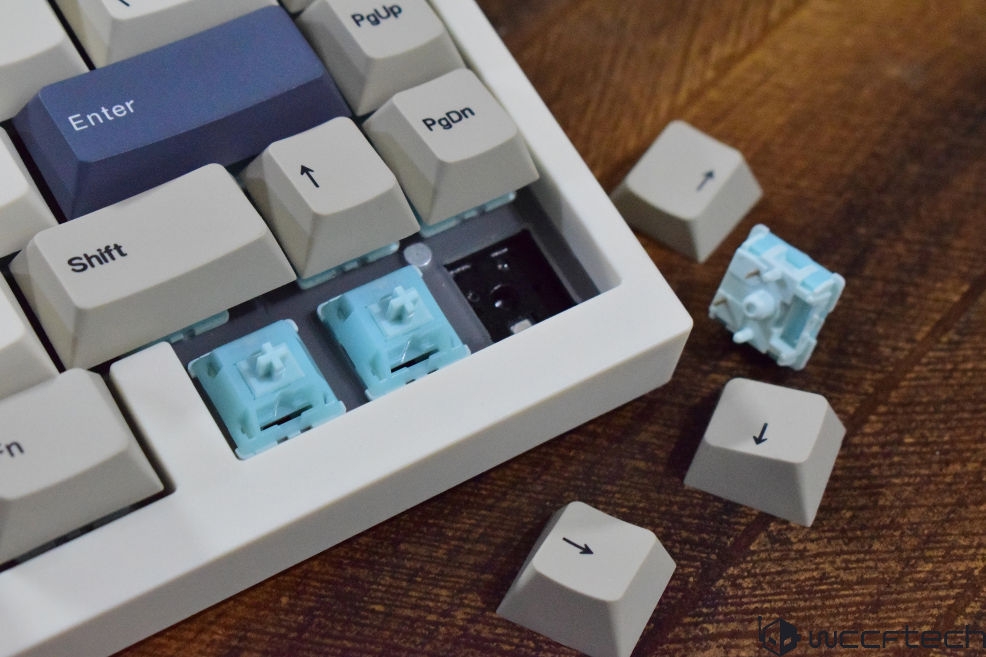 A mechanical keyboard with several detached gray keycaps exposes blue mechanical switches on a wooden surface.