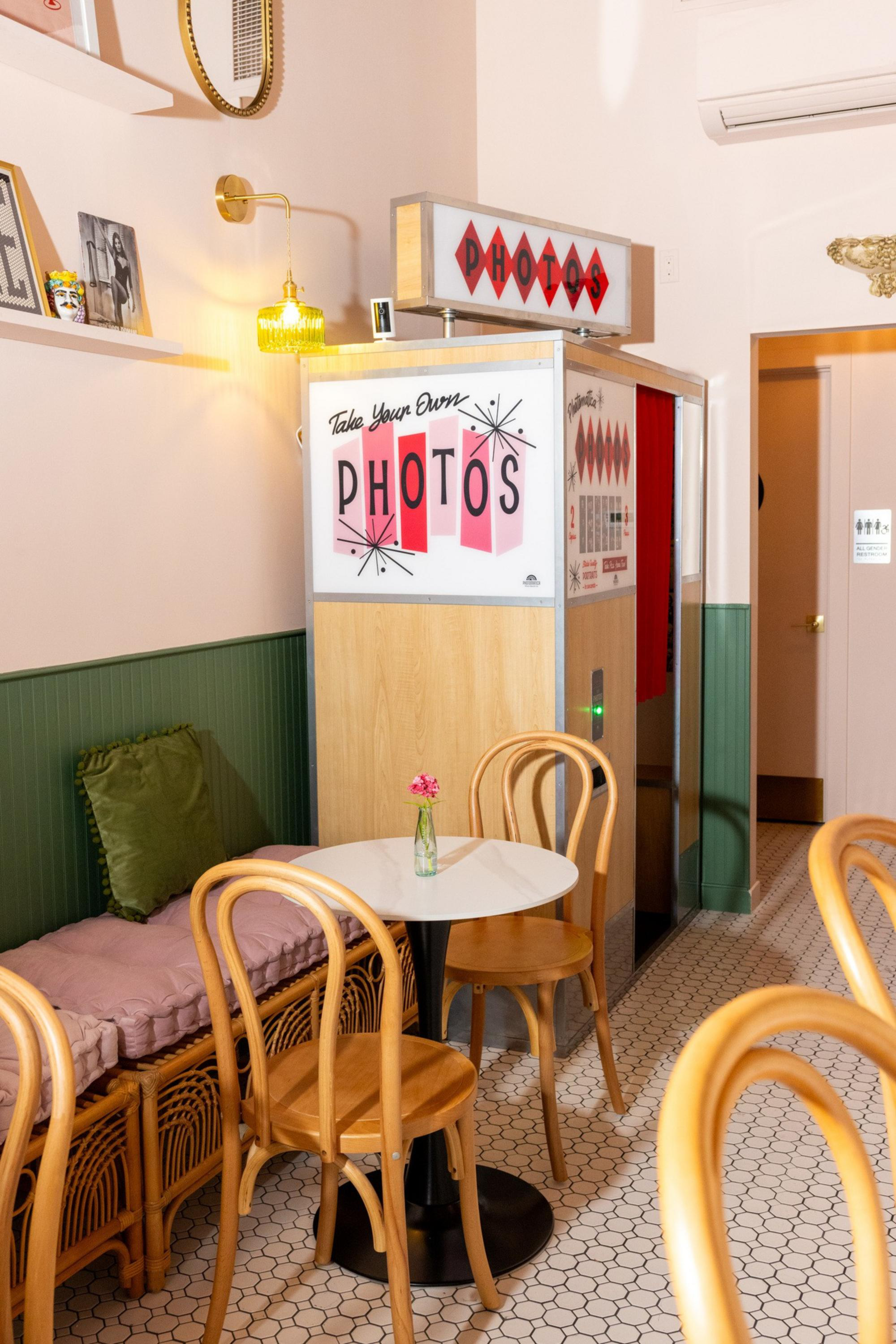 A cozy corner with a vintage photo booth, round white table, wooden chairs, cushioned wicker bench with green pillow, small flower vase, and tiled floor.