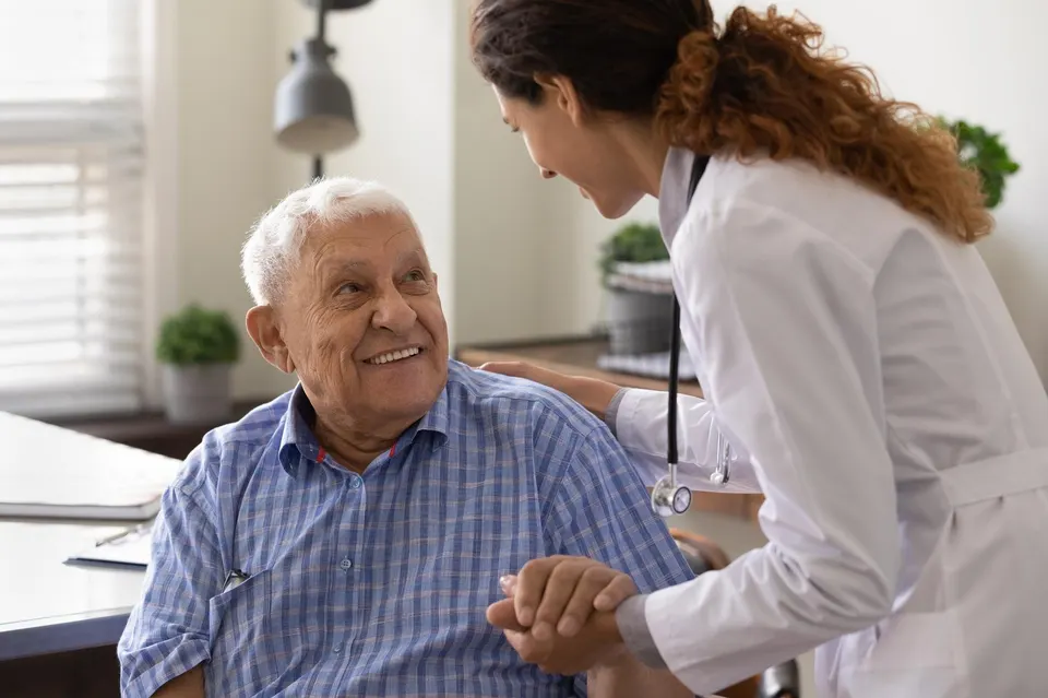 Close up caring nurse holding smiling mature male patient's hand at meeting in hospital.
