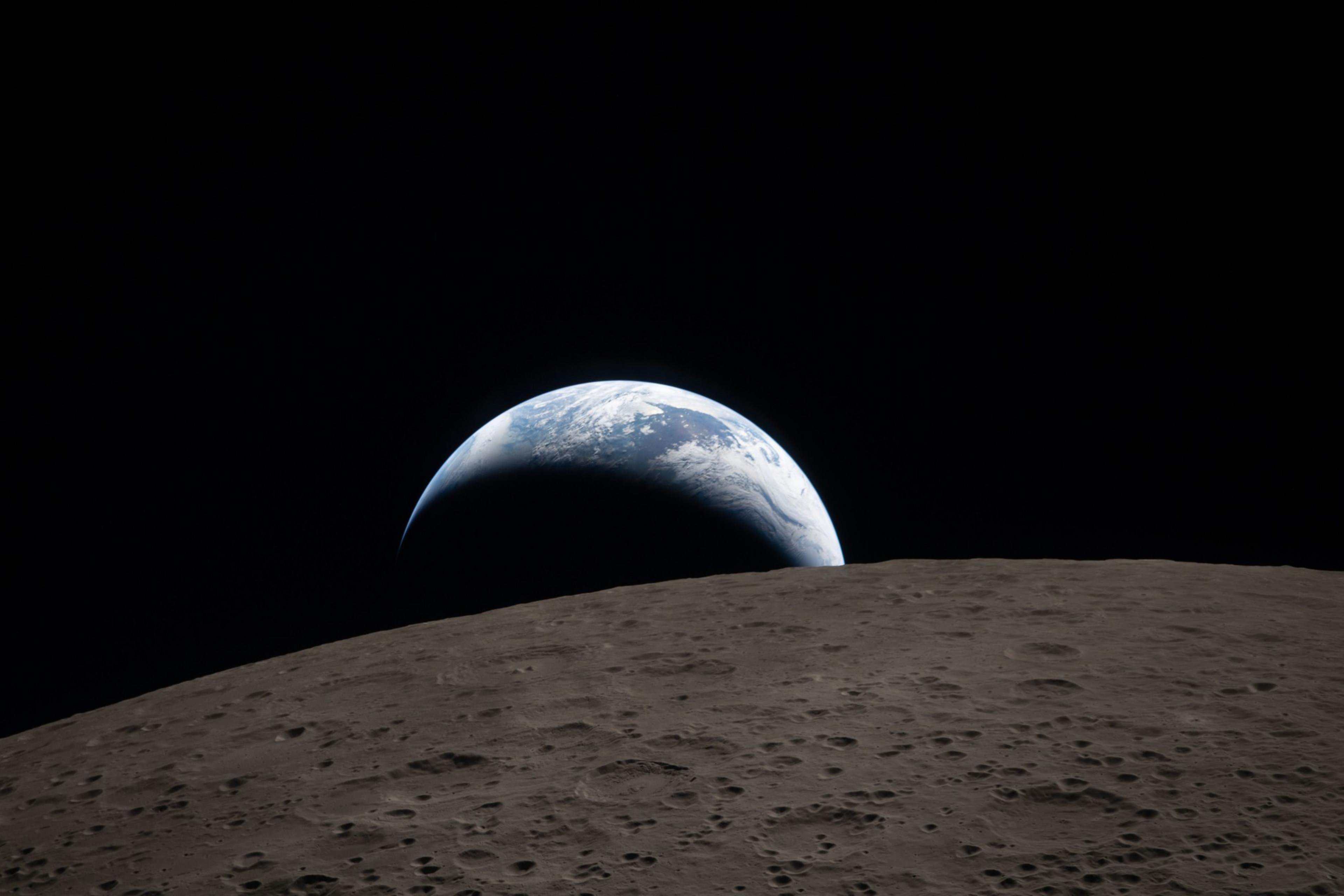 A partially illuminated Earth rises above the dark, cratered surface of the moon against the blackness of space.