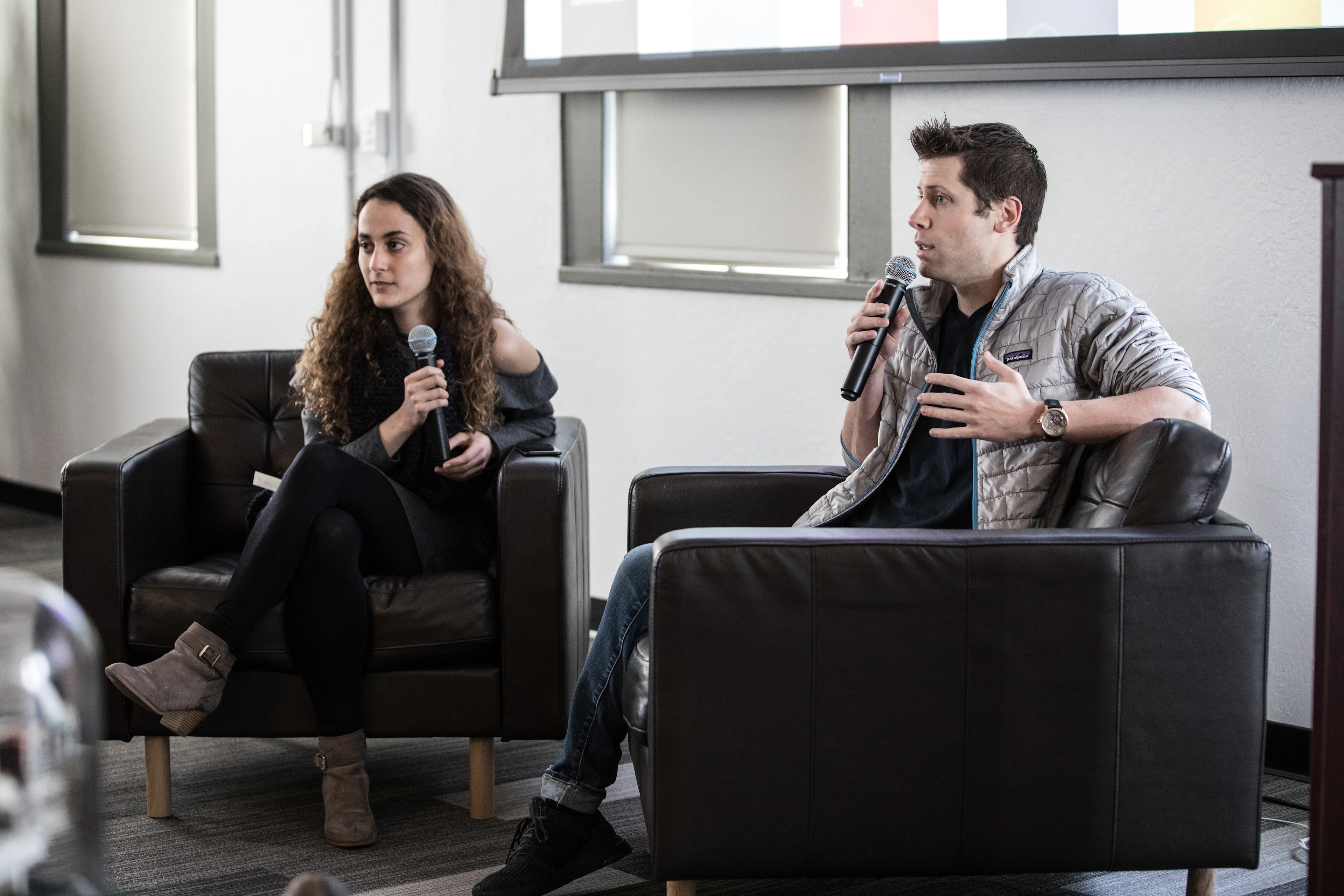 Two people seated in armchairs holding and talking into microphones